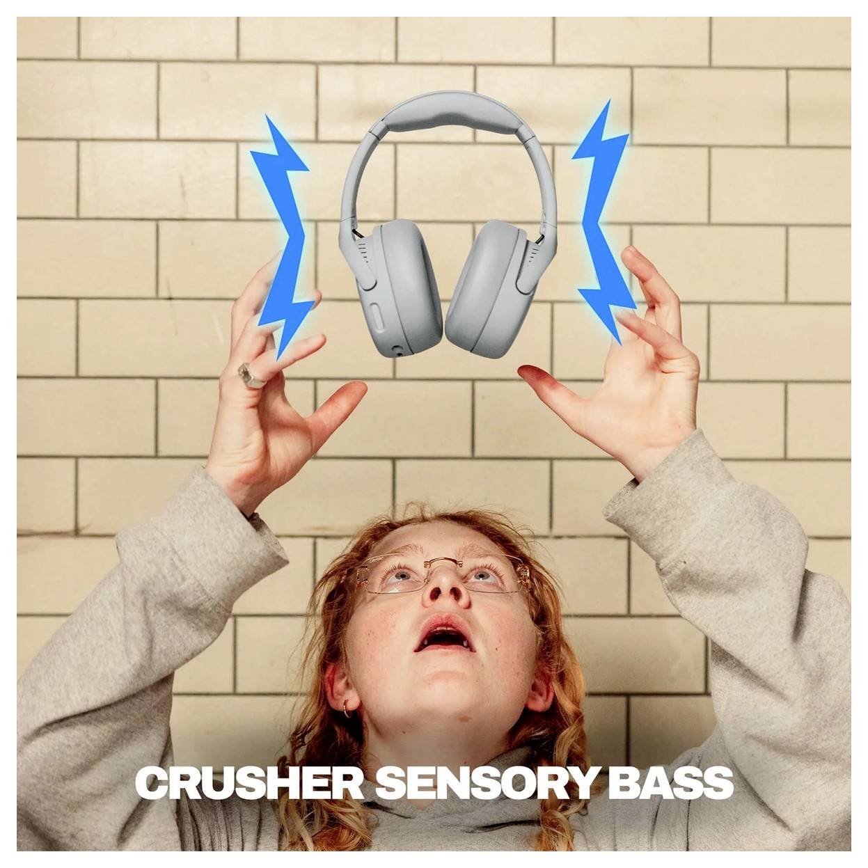 Oseba, ki sega navzgor proti lebdečim sivim slušalkom z modrimi streli; besedilo se glasi "CRUSHER SENSORY BASS".