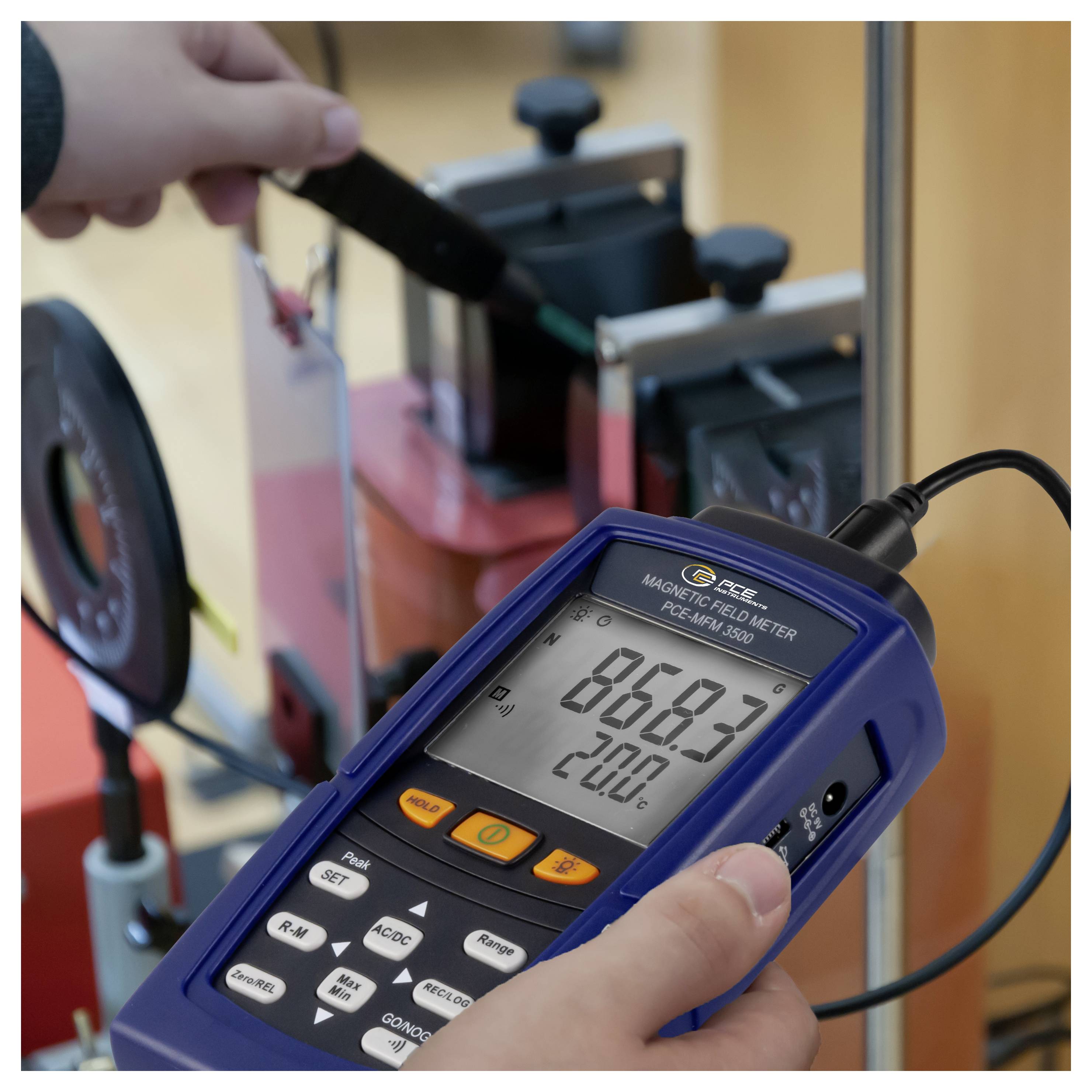 Oseba z ročnim merilcem magnetnega polja blizu laboratorijske opreme, ki meri in prikazuje odčitek 868,3 in 20,0°C na zaslonu.