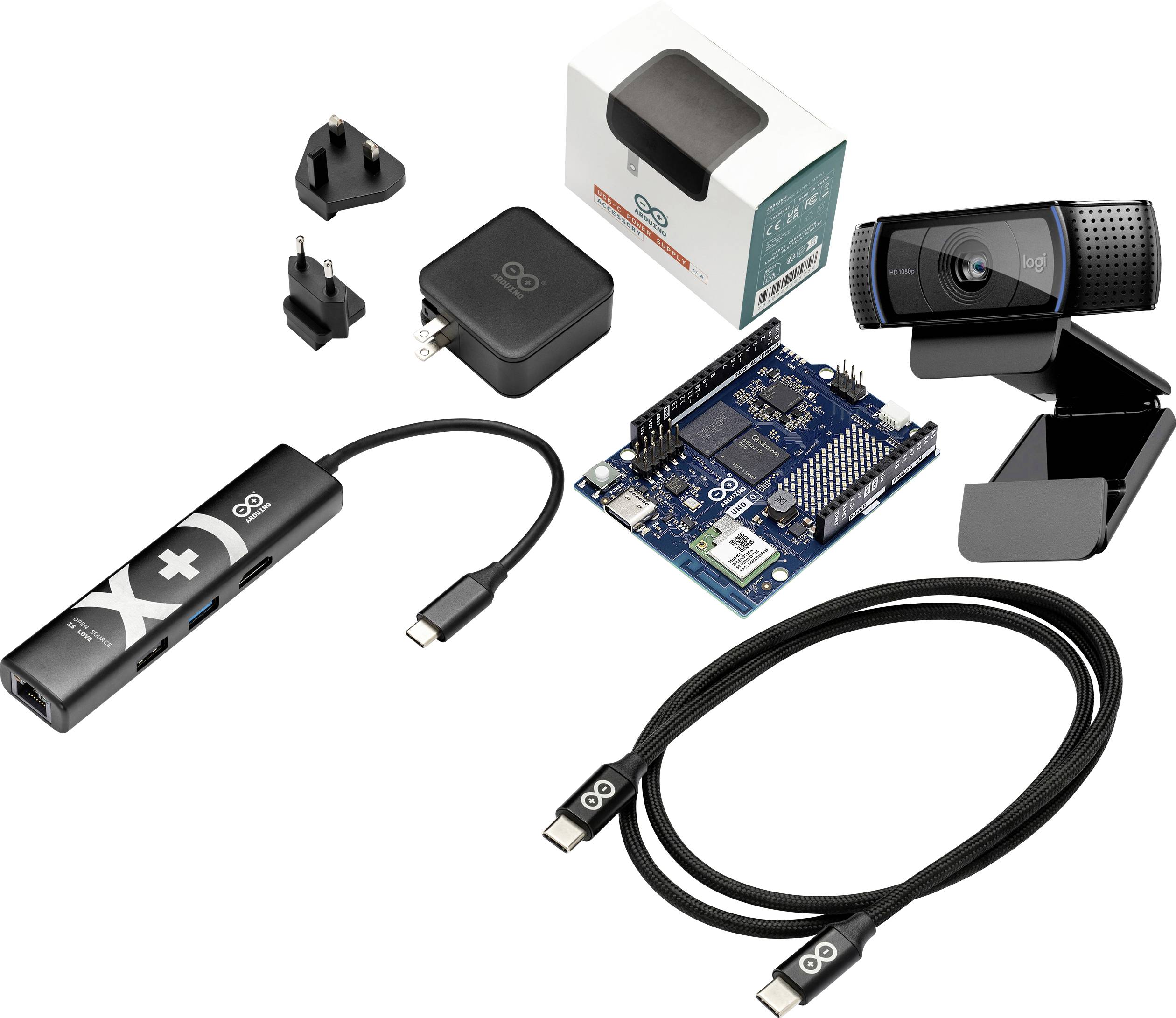 Komplet različnih elektronskih komponent: spletna kamera, plošča podobna Arduino-u, razdelilnik USB in kabli, napajalni adapter, prilagodni vtikači in trgovinska embalaža na belem ozadju.