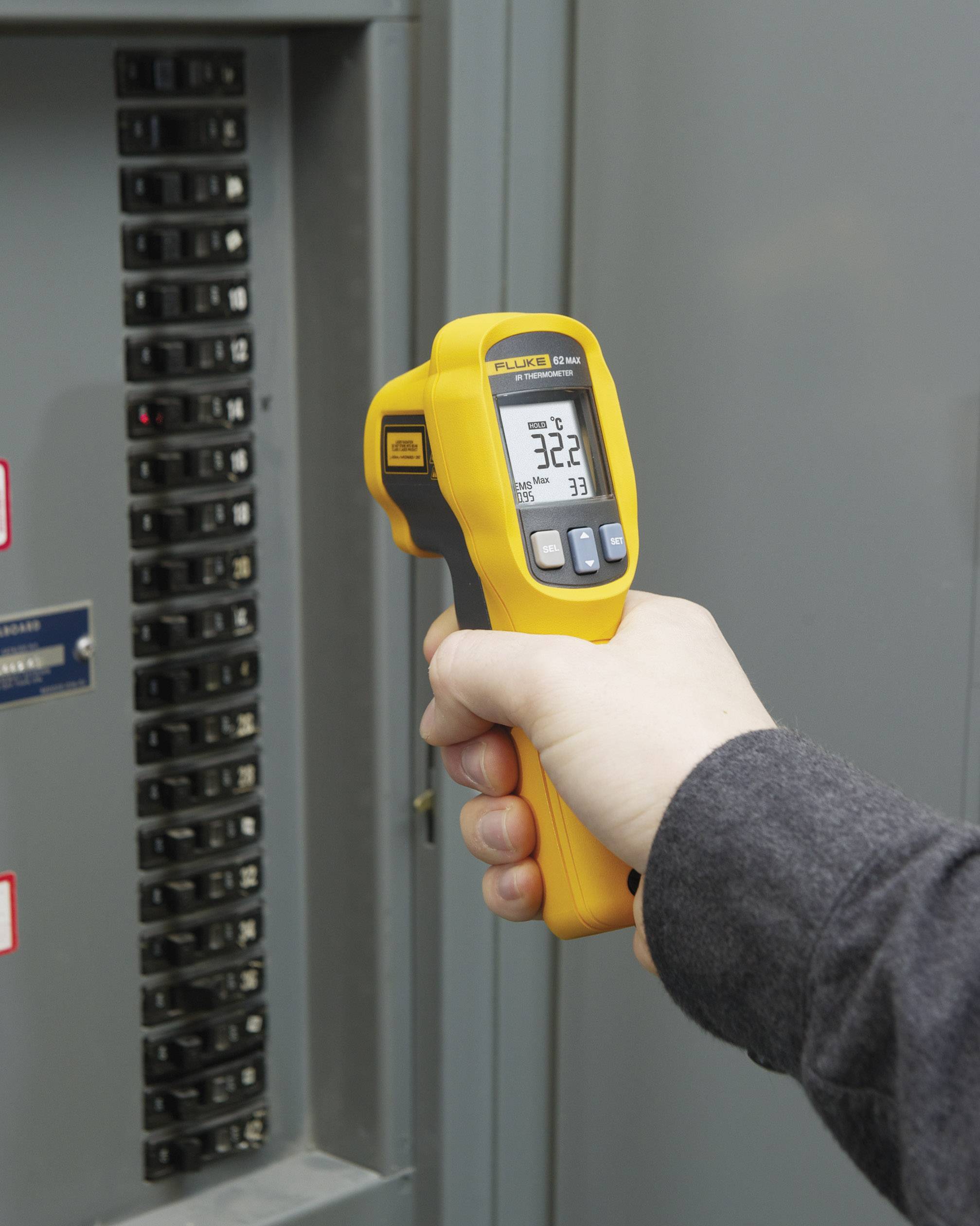 Roka drži rumeno infrardeče termometer pred električno omarico in na zaslonu prikazuje 32,2 stopinj Celzija.