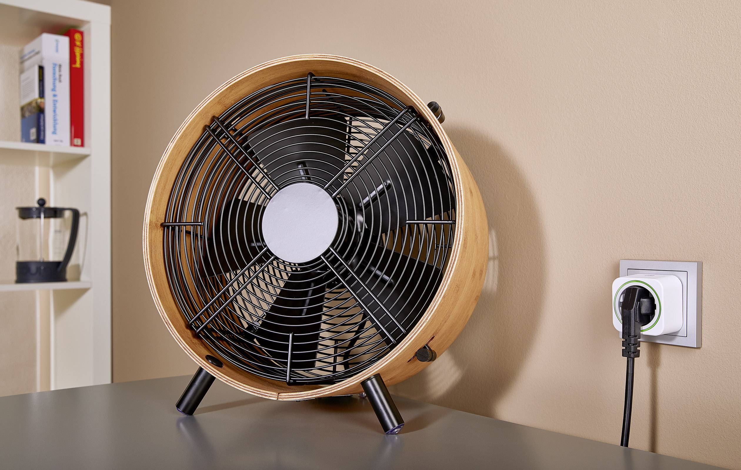 Retro namizni ventilator stoji na sivi mizi. Je priključen in pripravljen za uporabo. V ozadju je knjižna polica.