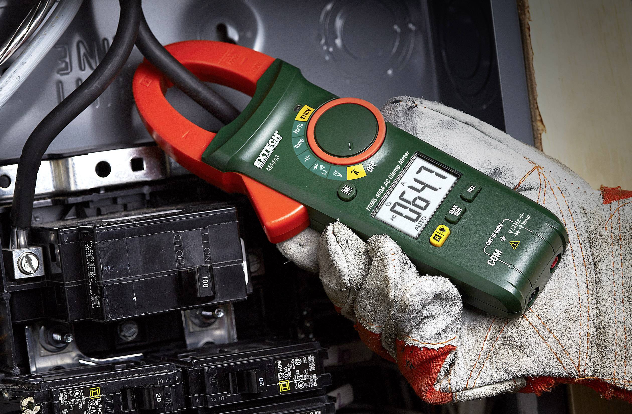 Oseba drži zeleni multimeter ob električni komponenti in meri napetost 164,7 voltov. Naprava prikazuje različne načine merjenja.