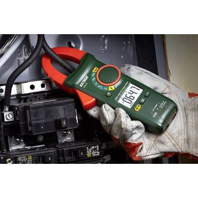 Oseba drži zeleni multimeter ob električni komponenti in meri napetost 164,7 voltov. Naprava prikazuje različne načine merjenja.