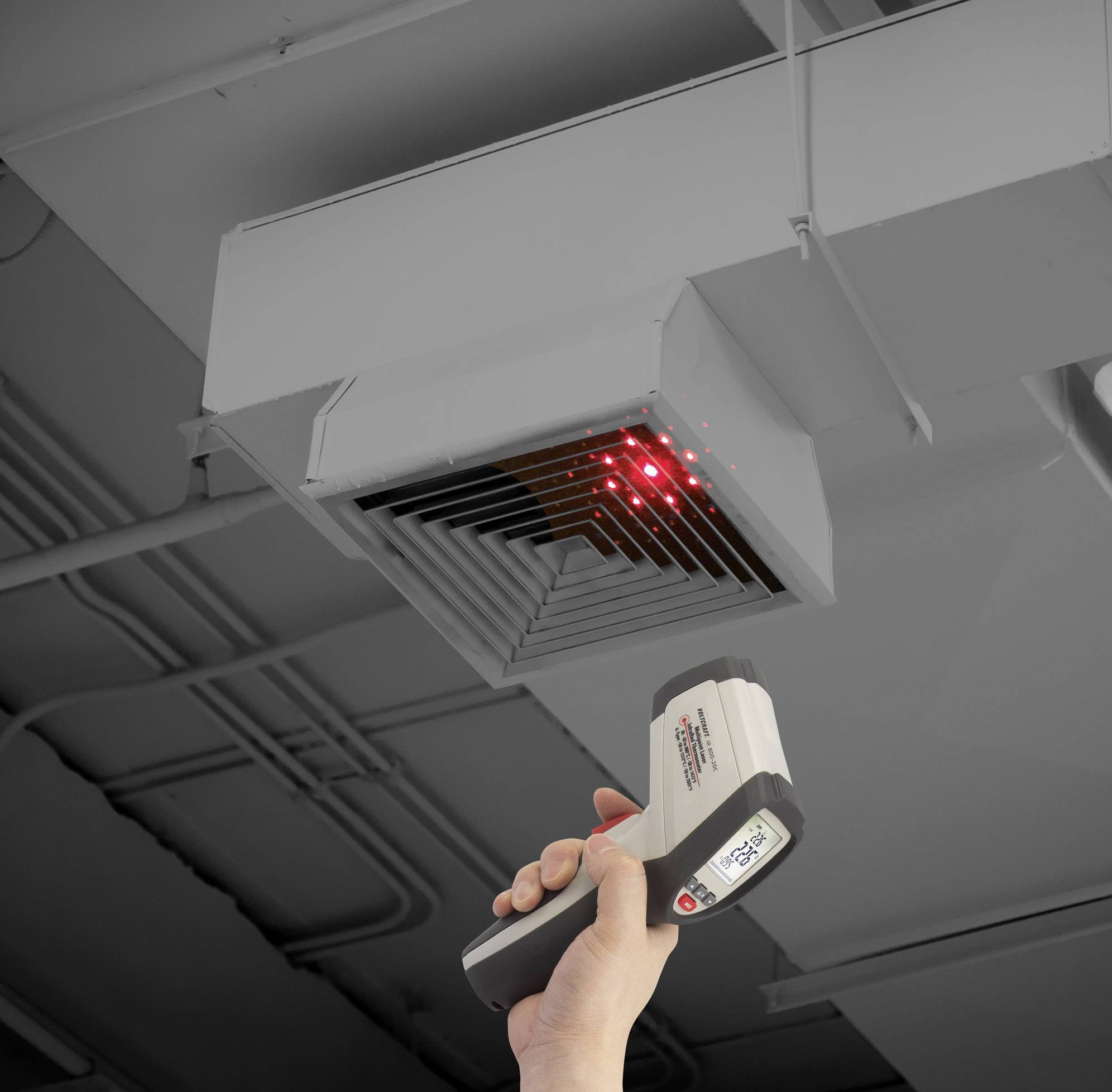 Oseba usmerja infrardeči termometer proti stropni prezračevalni rešetki, verjetno za merjenje temperature.