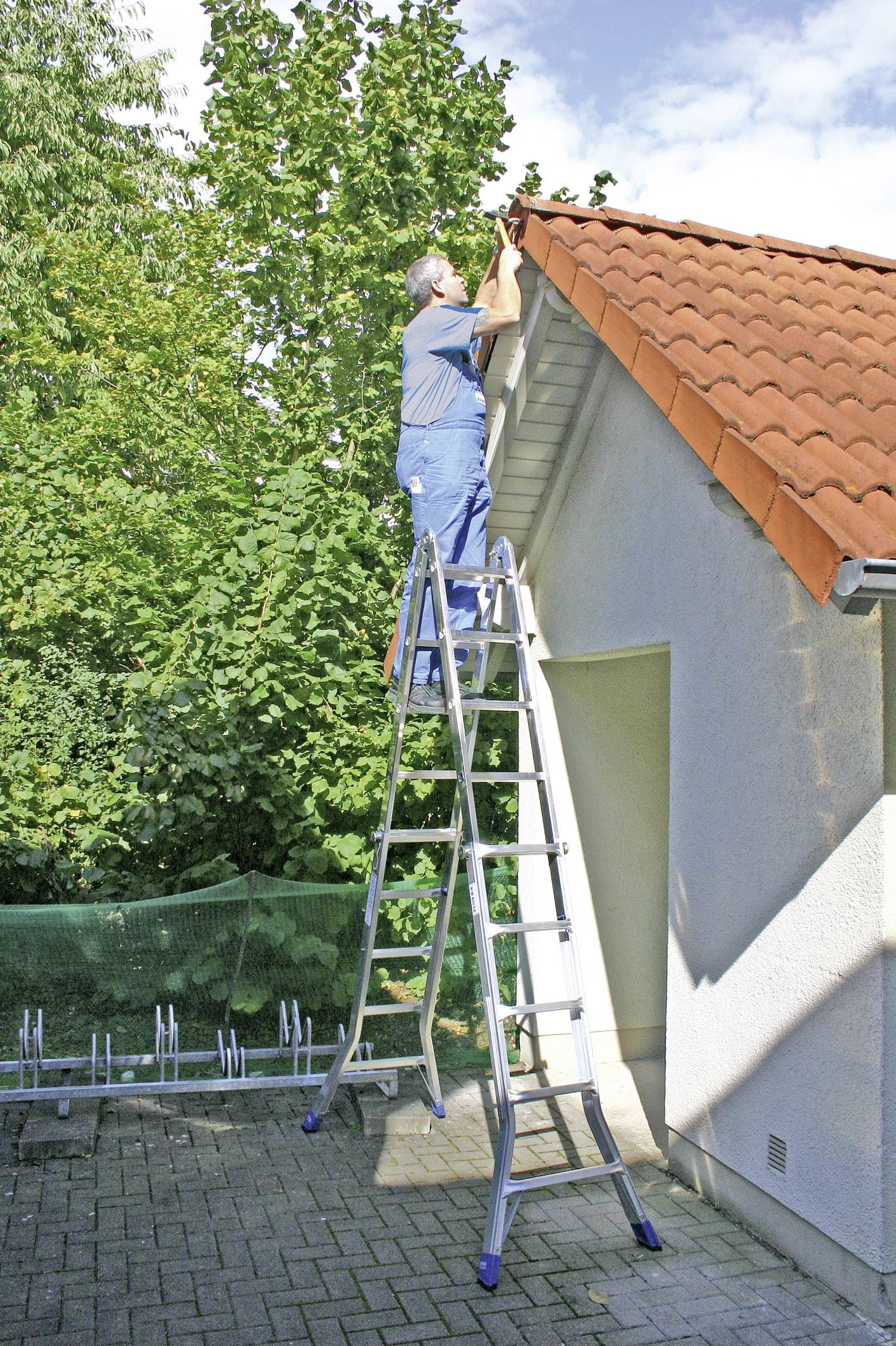 Oseba stoji na visoki lestvi in popravlja streho hiše zunaj pri dnevni svetlobi, obkrožena z zelenimi drevesi.