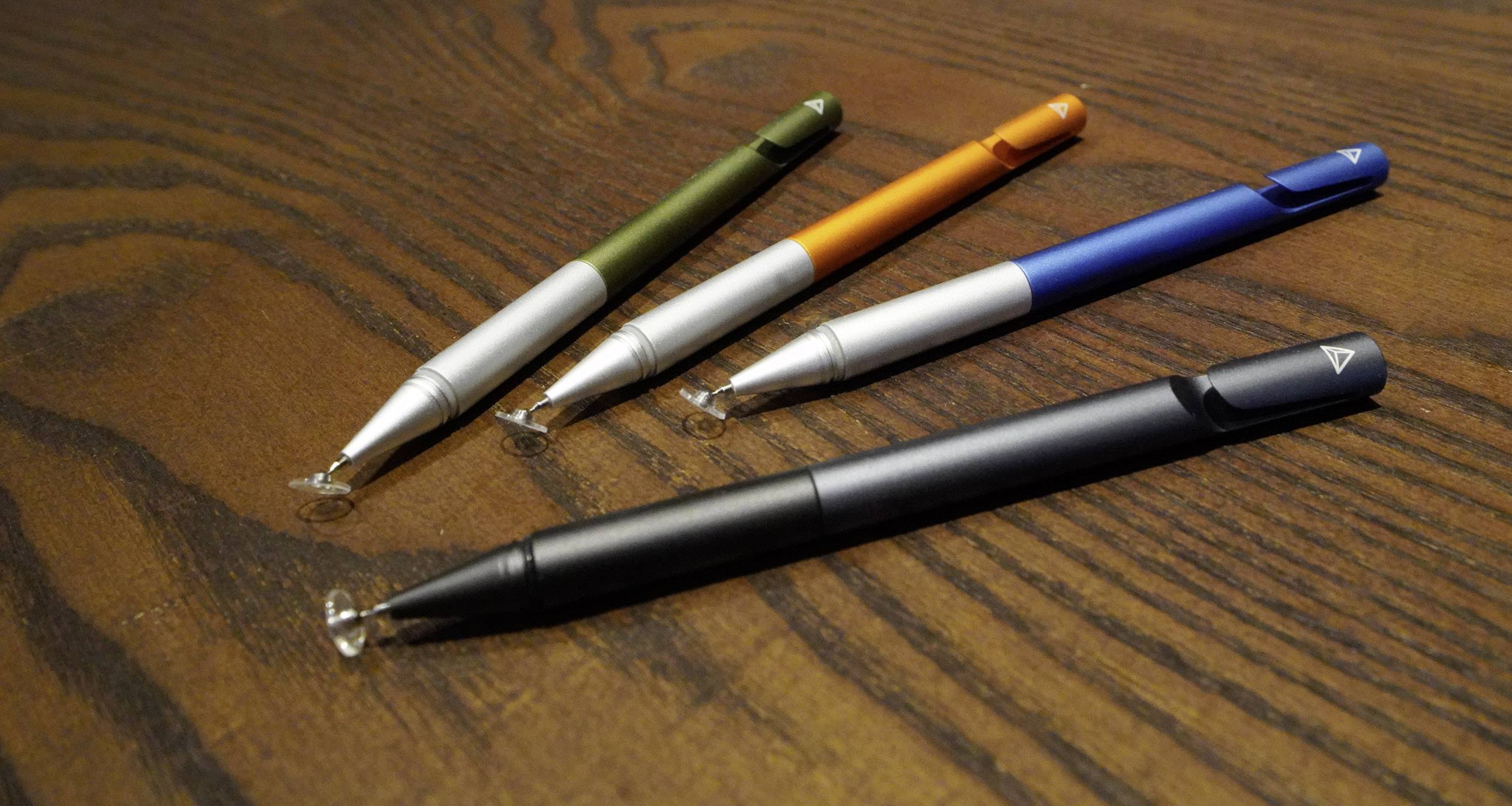Štirje svinčniki stylus različnih barv (zelena, oranžna, modra, črna) ležijo na leseni mizi. Imajo prosojne, okrogle konice.