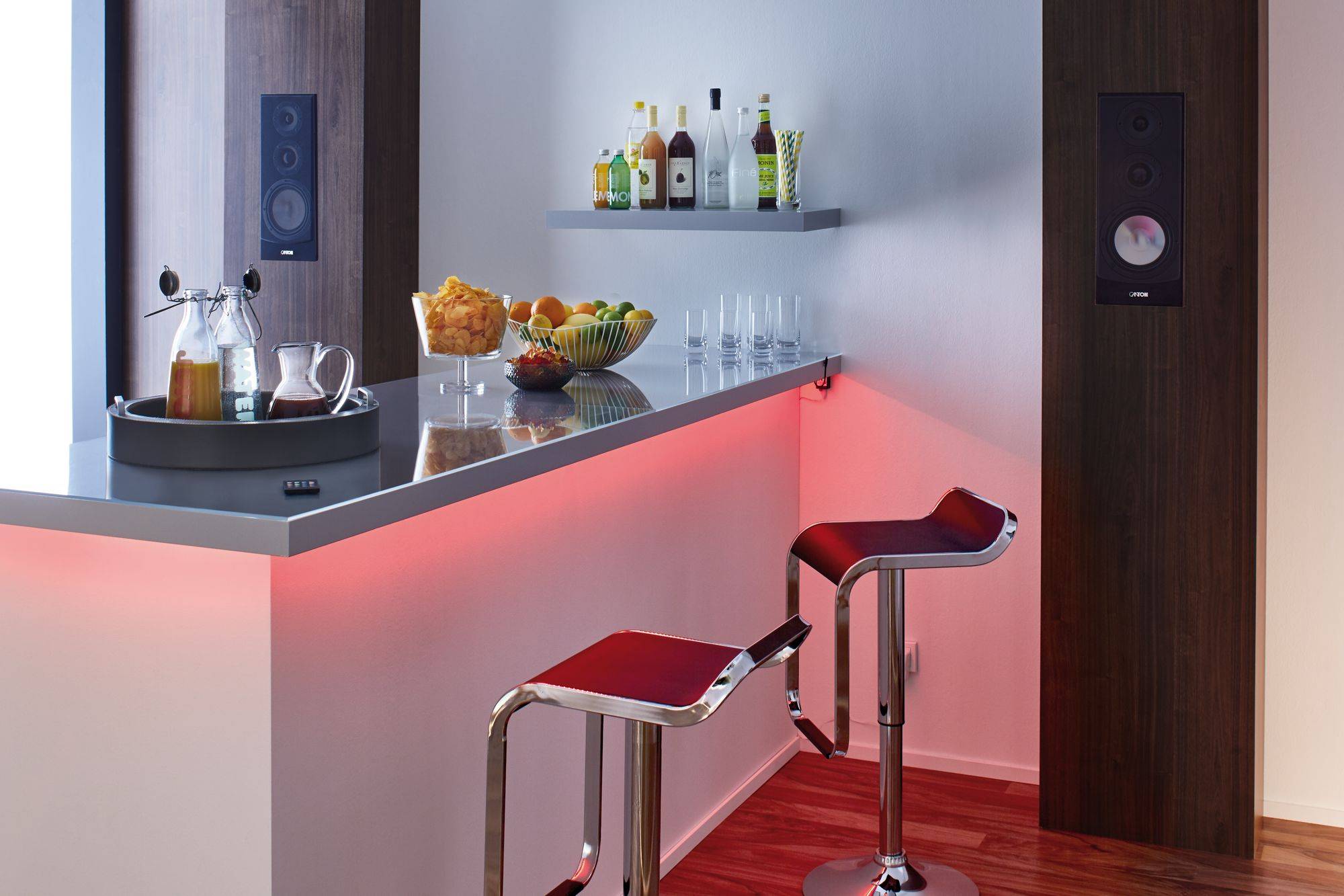 Moderna domača bar z napitki na pultu, vključno z sokom in žganimi pijačami. Pred njim sta dva rdeča barska stolčka. Subtilna osvetlitev.