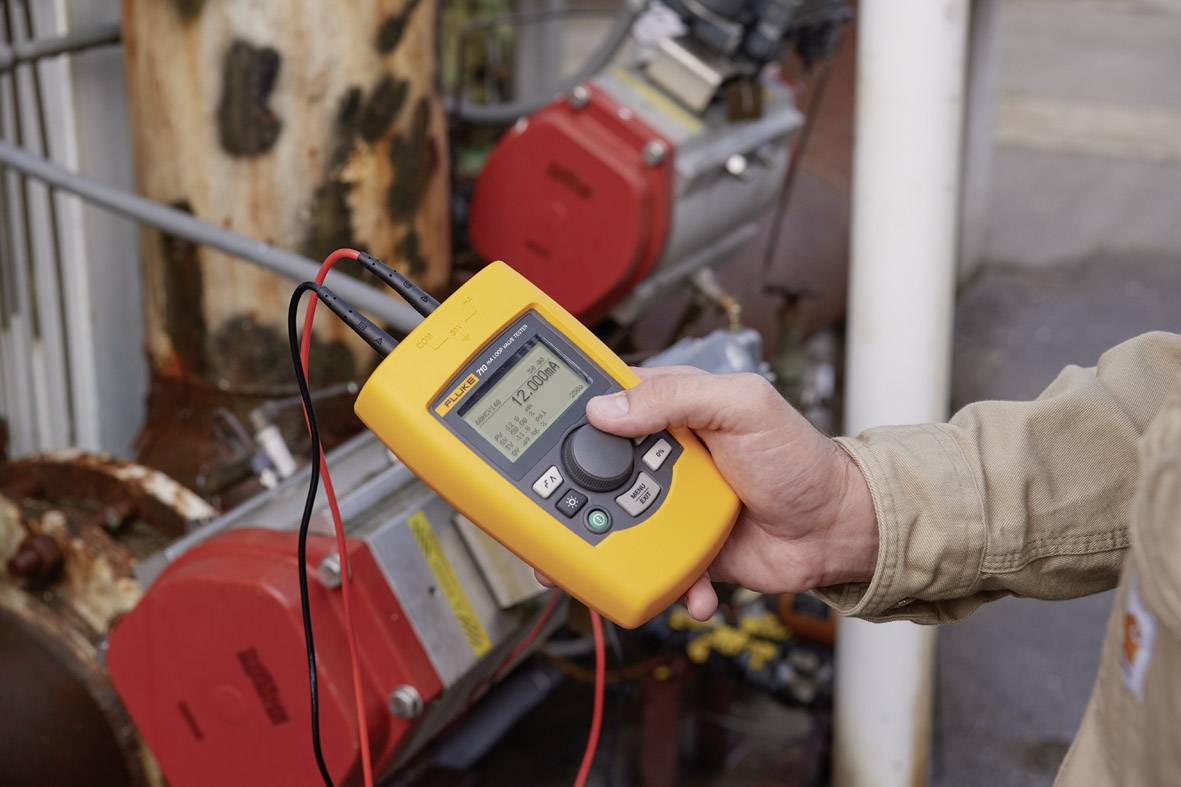 Oseba drži rumeno digitalno merilno napravo, ki prikazuje električne vrednosti. V ozadju so vidni industrijski stroji.