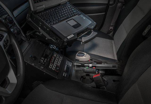 Prenosni računalnik in radijske postaje, nameščene v policijskem vozilu; prikazuje tehnologije za komunikacijo in dostop do podatkov med patruljiranjem.