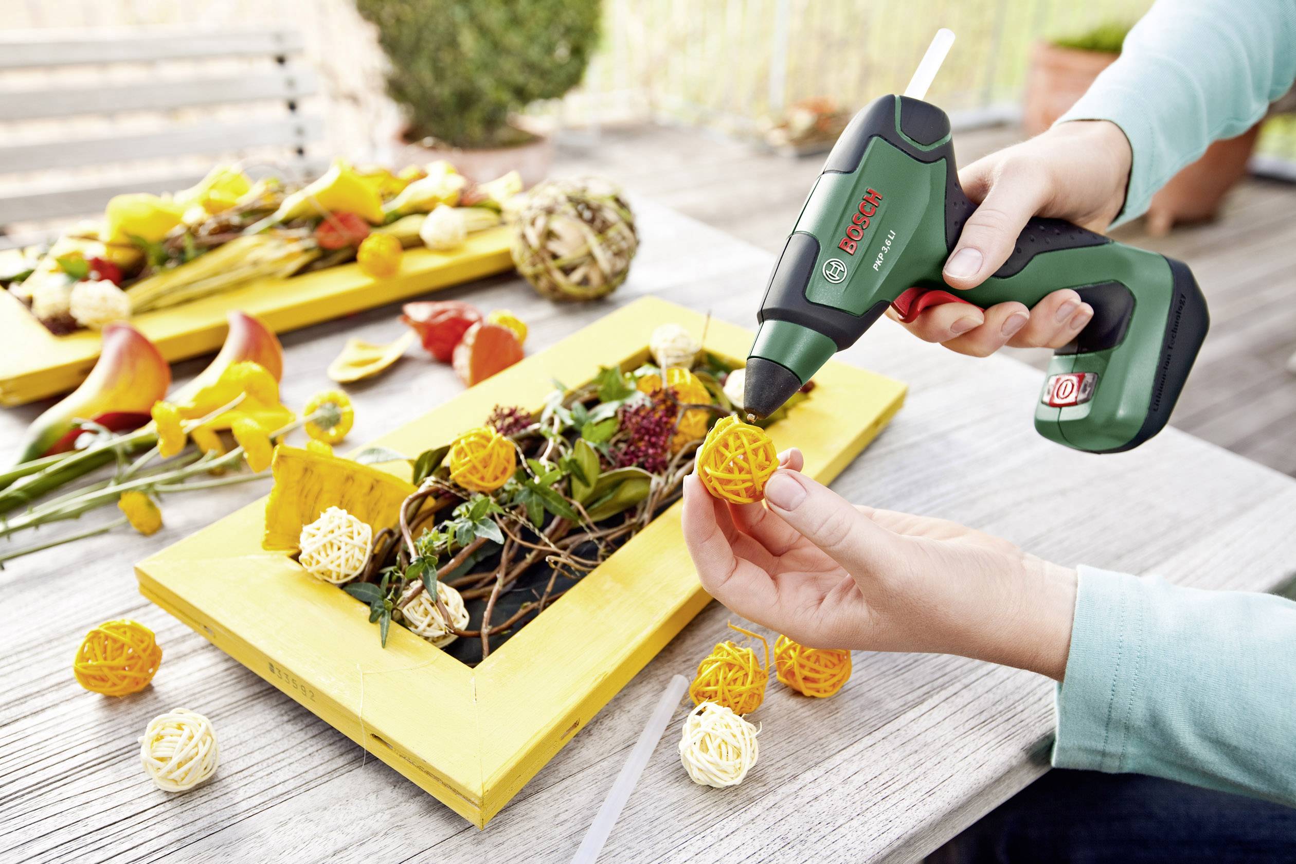 Oseba uporablja vročo lepilno pištolo za ustvarjanje dekoracije DIY z rumenimi cvetovi in suhim materialom na rumenem okvirju.