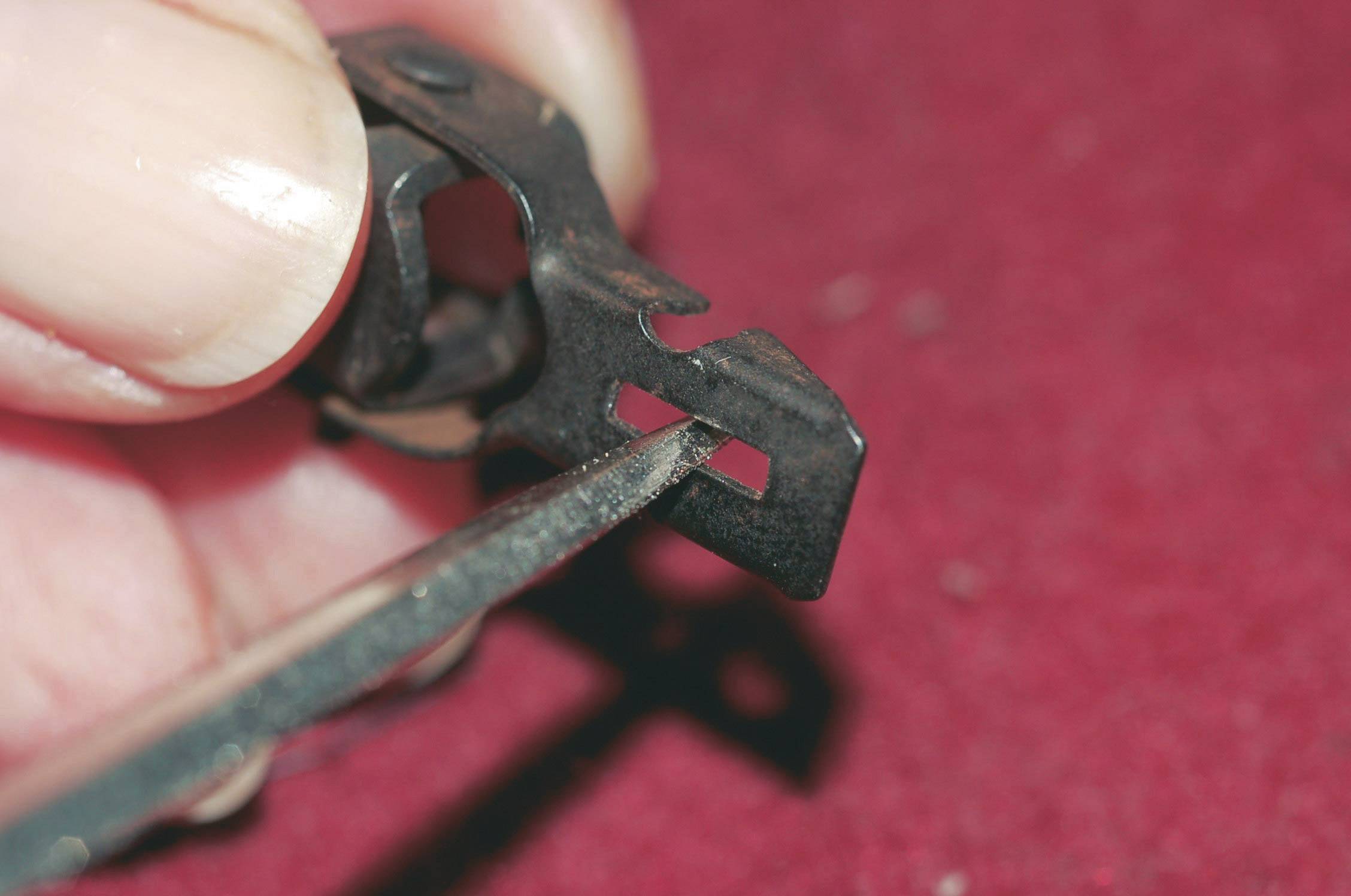 Ena roka drži ozko kovinsko orodje ob majhen črn strojni del z pravokotnimi izrezi na rdeči podlagi.