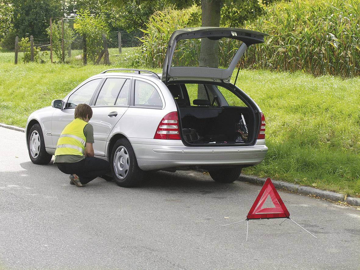 Srebrn avto z odprtim prtljažnikom stoji ob robu ceste. Oseba v odsevnem jopiču kleči poleg avtomobila. Postavljeni so opozorilni trikotnik.