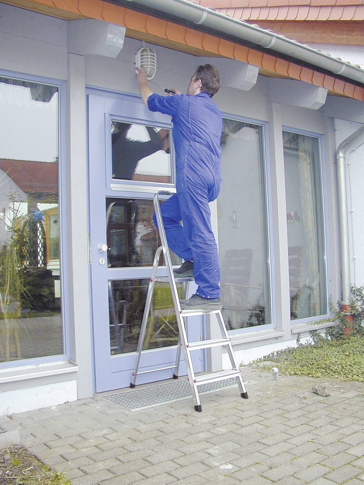 Moški v modri delovni opremi stoji na lestvi in popravlja zunanjo svetilko nad vhodnimi vrati stavbe.