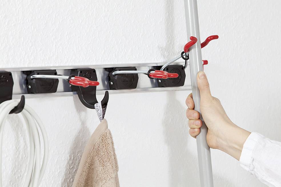 Roka drži držalo metle, pritrjeno v stenski nosilec z več rdečimi sponkami. Poleg visi brisača in kabel.