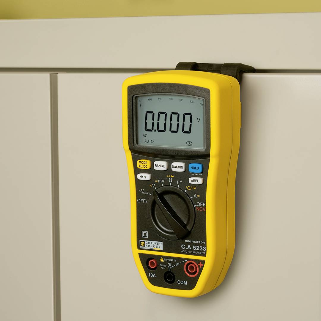 Ett gult digitalt multimeter hänger på ett vitt skåp. Displayen visar '0,000 V'. Olika mätfunktioner kan väljas.