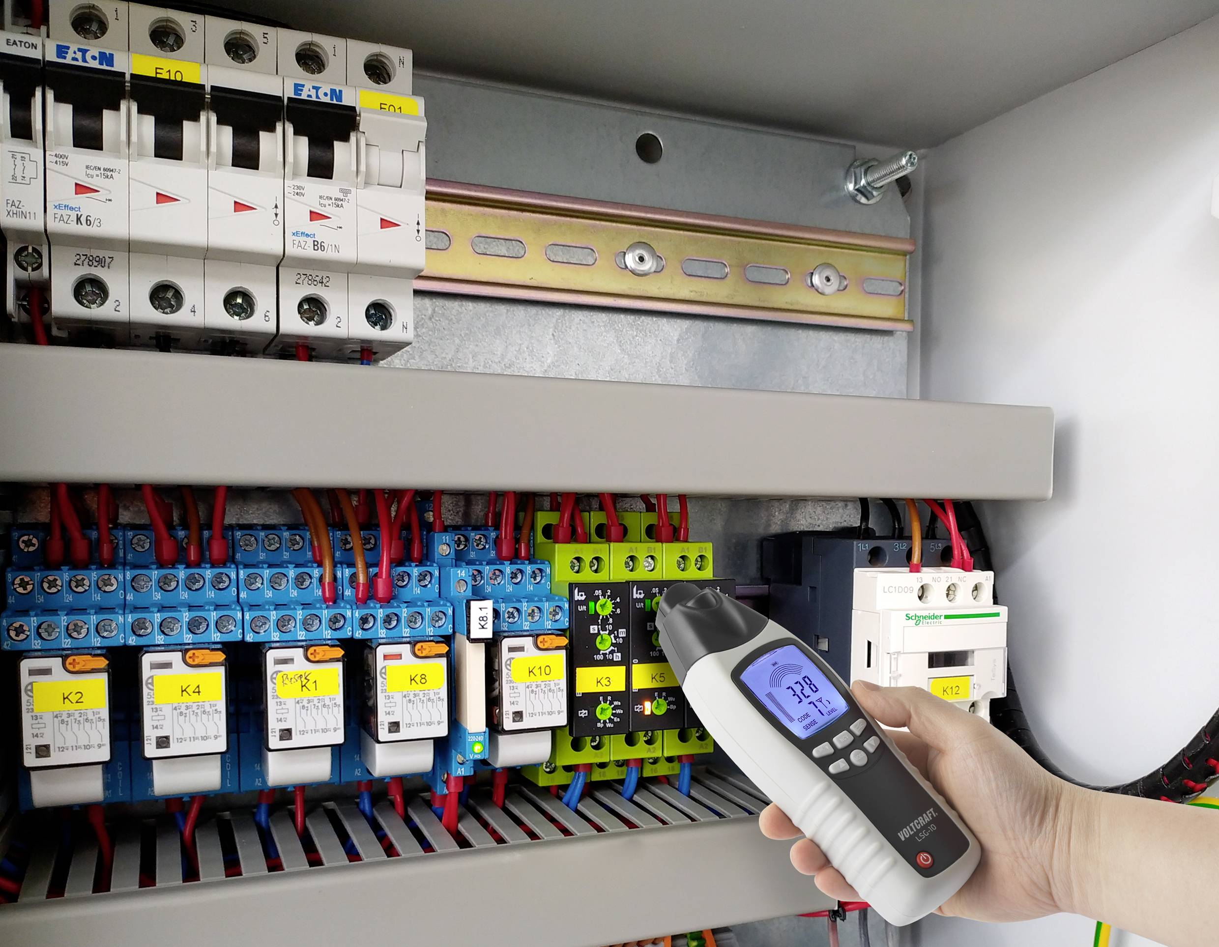En hand håller ett multimeter som mäter spänningen i ett öppet elskåp. Olika switchar och kabeldragningar syns.