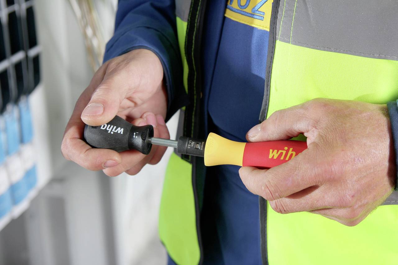 En person håller två skruvmejslar med svarta och röda handtag i händerna. Bakgrunden är oskarp med teknisk utrustning.