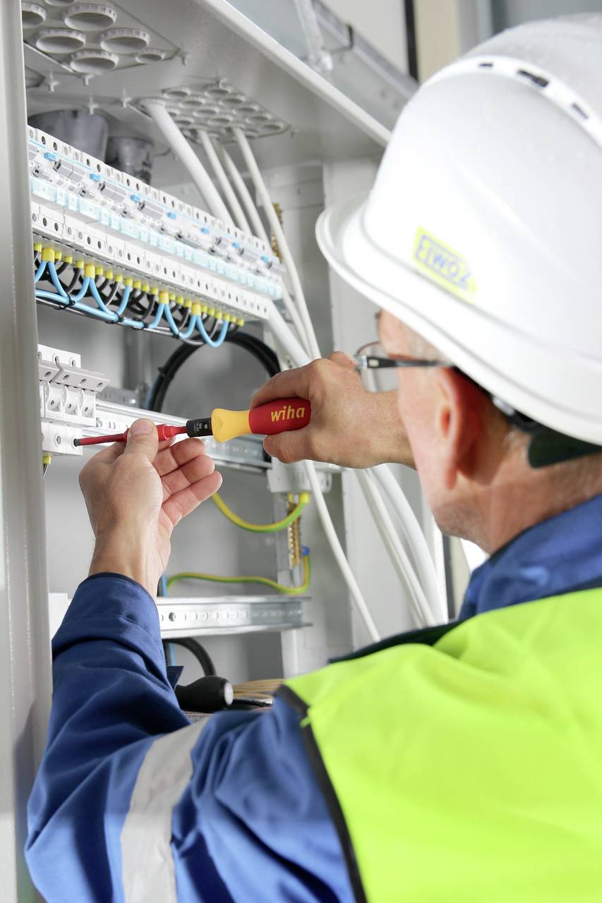 En elektriker med skyddshjälm och varselväst arbetar vid en elcentral och använder en skruvmejsel för att göra anslutningar.