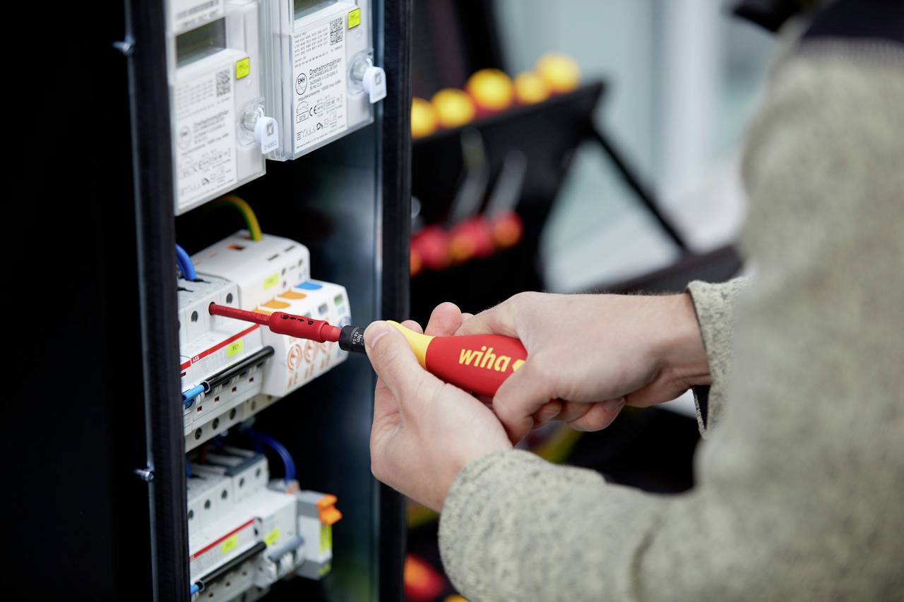 En person använder en skruvmejsel för att arbeta vid en elektrisk kopplingscentral. I bakgrunden syns verktyg.