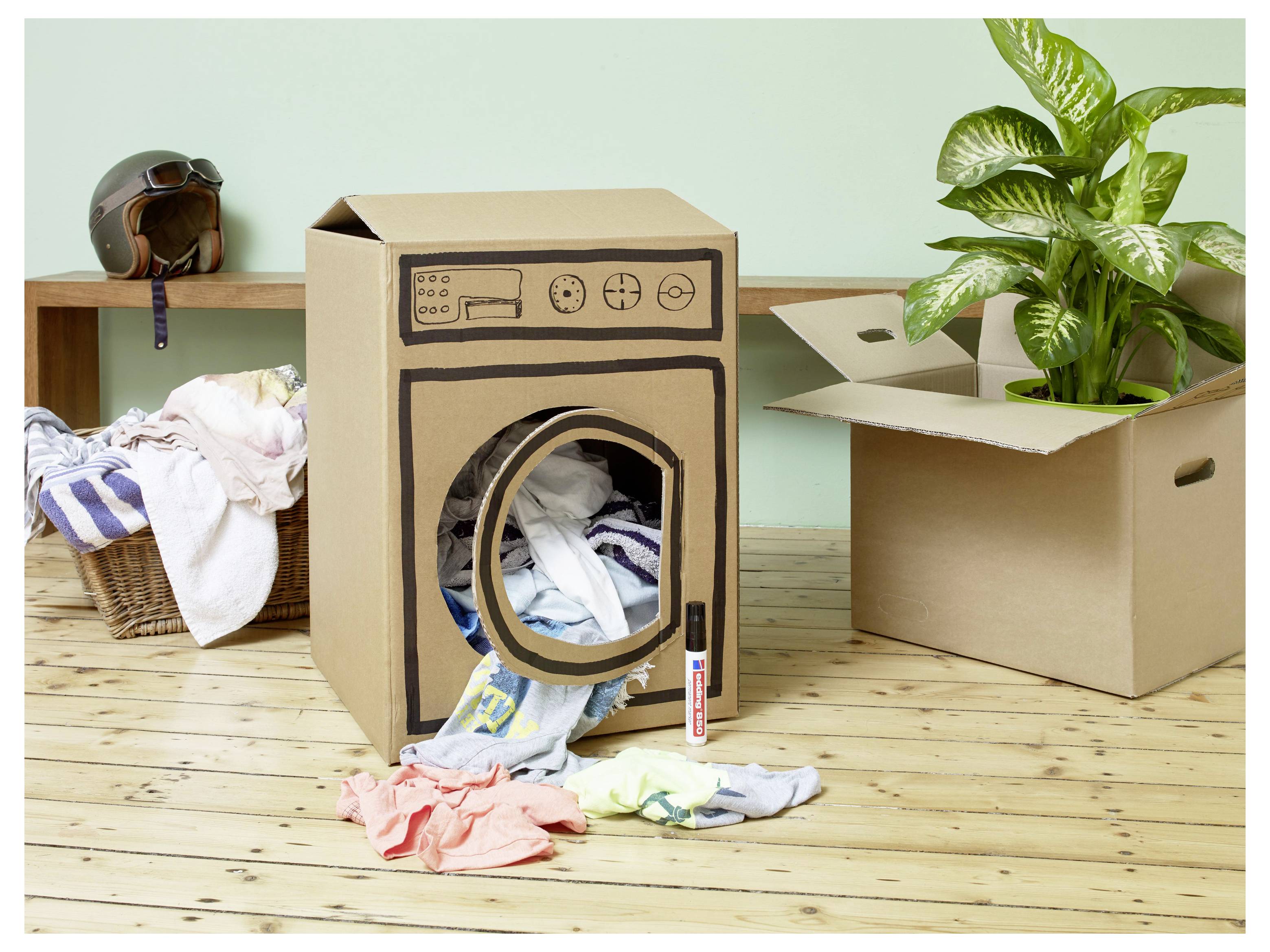 Kartongask dekorerad som en tvättmaskin med kläder som spiller ut, bredvid en tvättpkorg, en öppen flyttask och en krukväxt i ett rum.