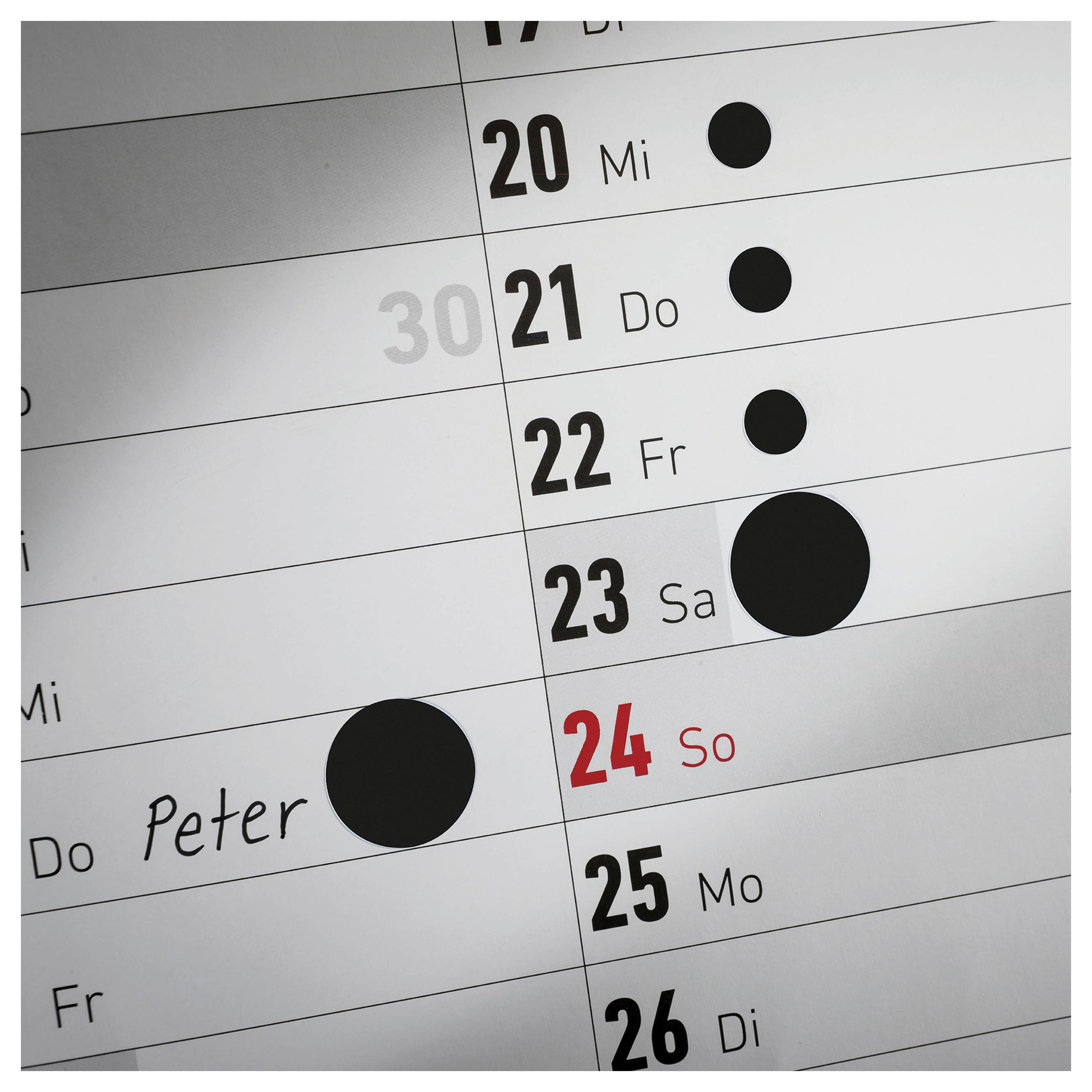 Närbild av en väggkalender med dagarna 20 till 26; den 24:e är markerad i rött som söndag, flera svarta punkter markerar tidpunkter, en gång med namnet "Peter".