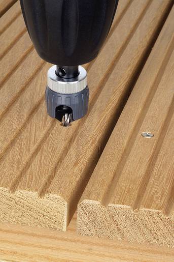 Närbild av en borr som borrar in i en träplanka. Plankan har en räfflad yta och ligger på en arbänk.