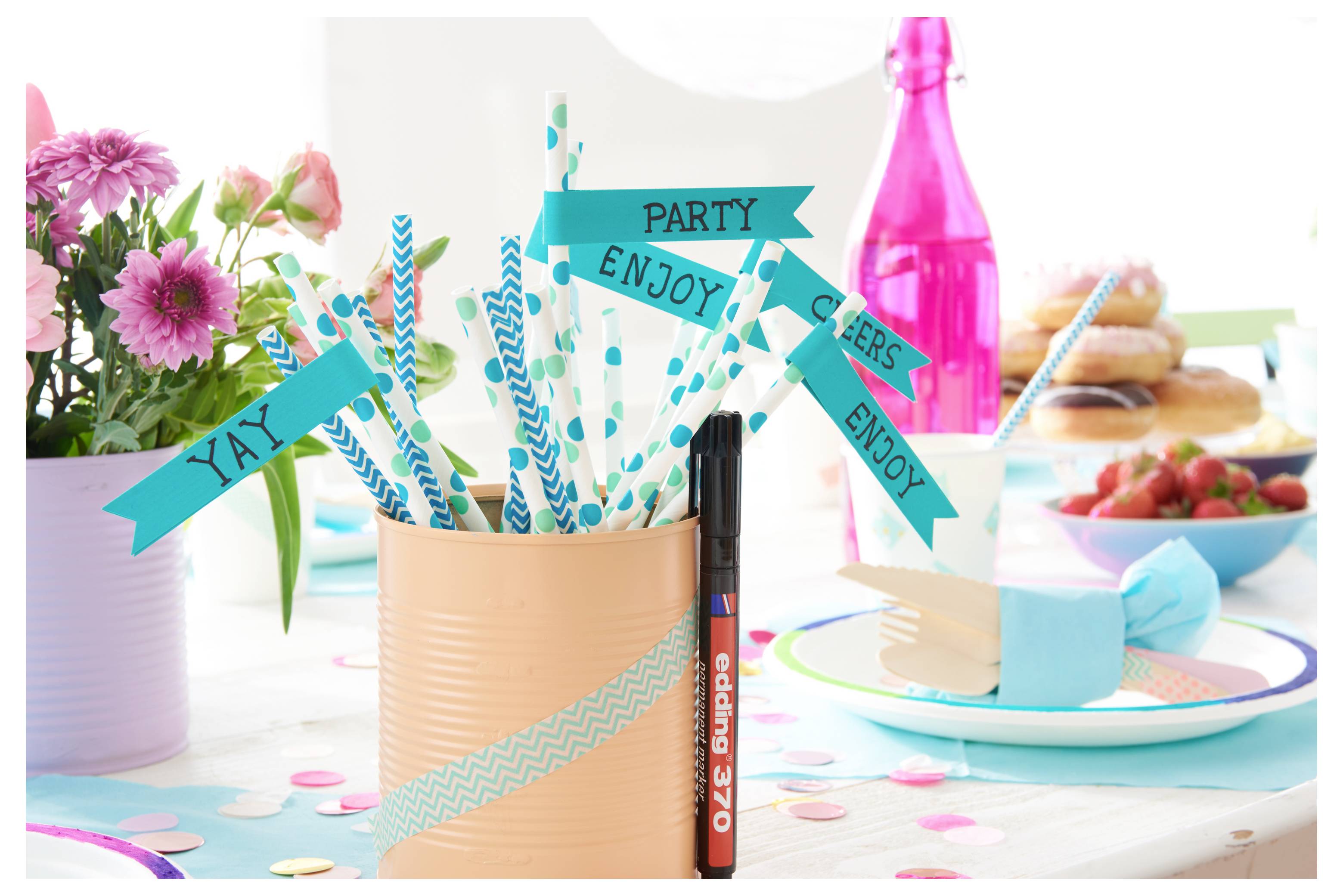 Partybordsinställning med teal-randiga sugrör och banderoller med texten "YAY", "PARTY", "ENJOY" i en persikofärgad kopp, plus efterrätter, blommor och en rosa flaska.