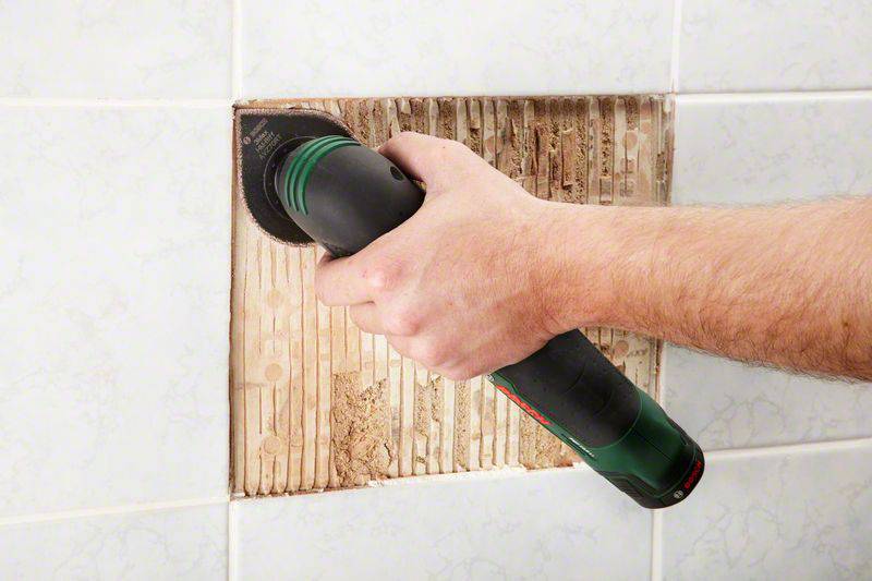 En person tar bort kakel från en vägg med ett elverktyg. Delar av det underliggande materialet är synligt.