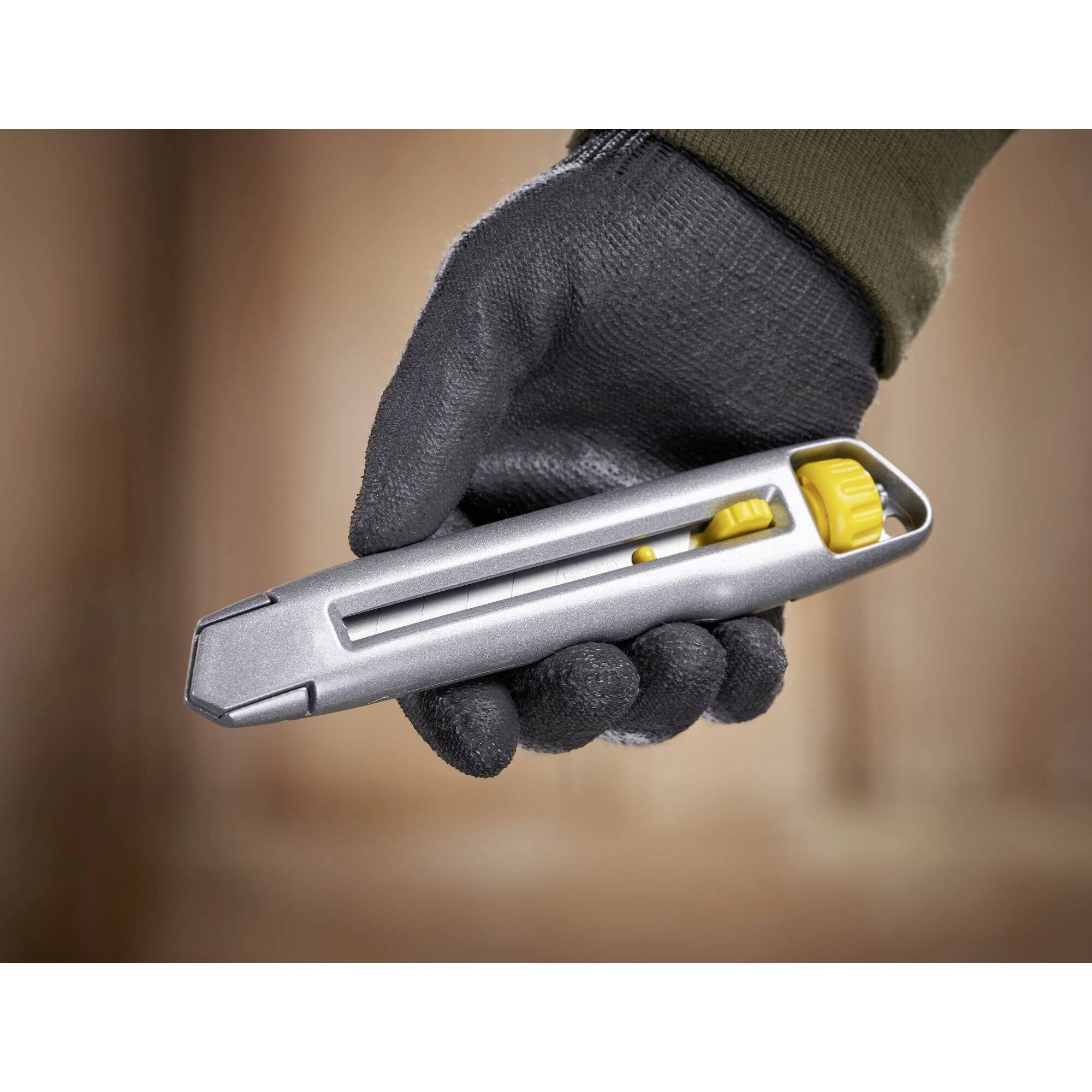 En handseklädd hand håller ett grått universalkniv med gul skjutreglage, redo att skära.