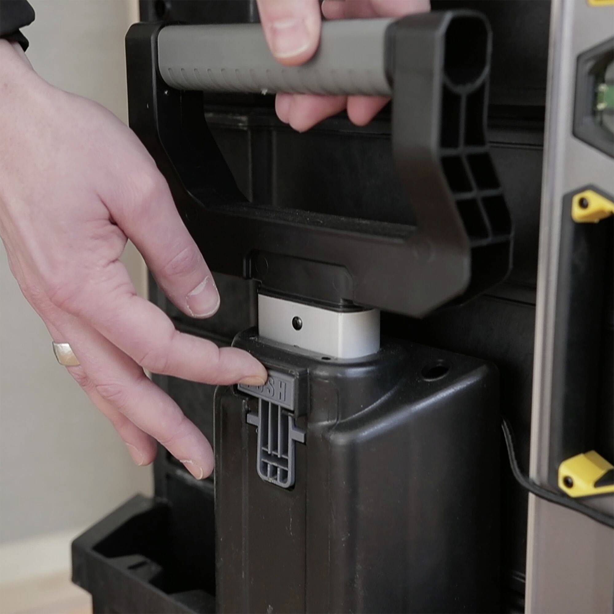 En person laddar batteriet till en elektrisk rullstol genom att sätta in det i laddaren. Händer syns nära enheten.