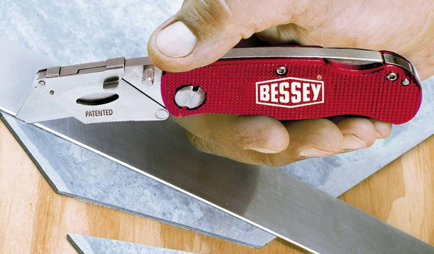 En hand håller en röd fällkniv med texten 'Bessey'. Trapetsblad och metalldelar ligger på ett träunderlag.
