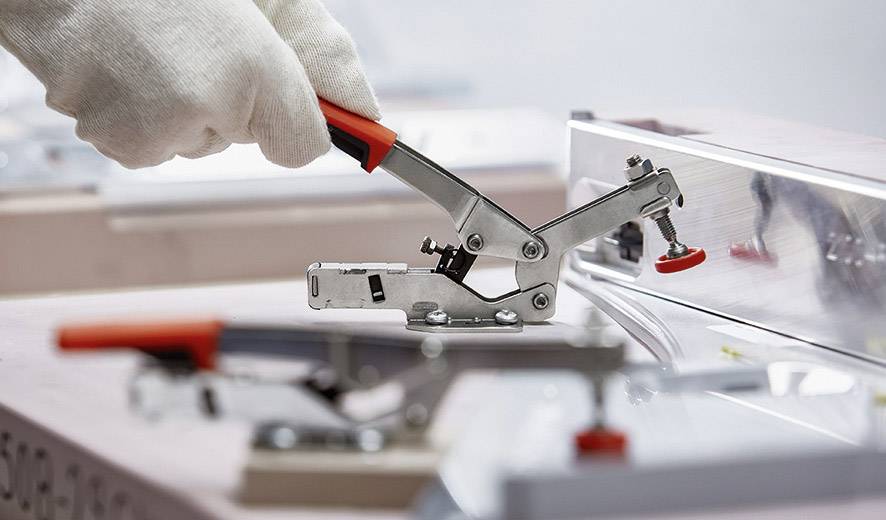 En handskklädd hand manövrerar ett metallspännverktyg med röd handtag, monterat på en maskin.