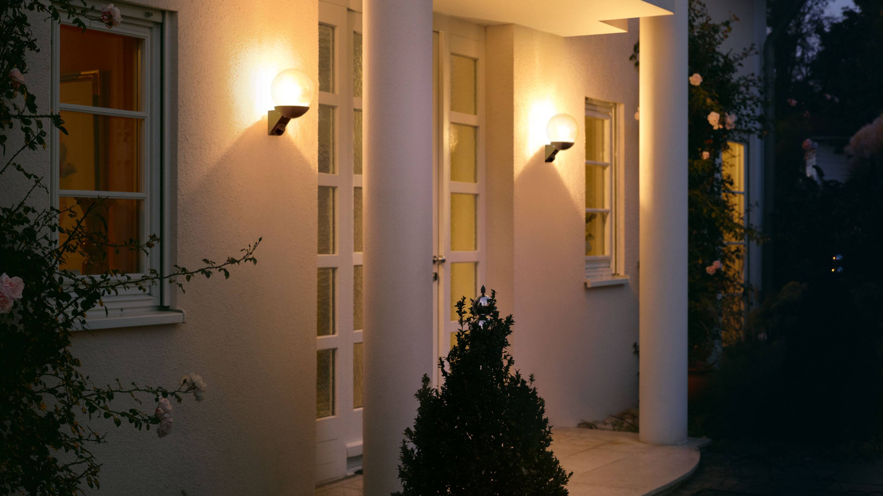 Entrén till ett hus med upplysta väggsconce, två stora pelare och en glasad dörr, omgiven av växter.
