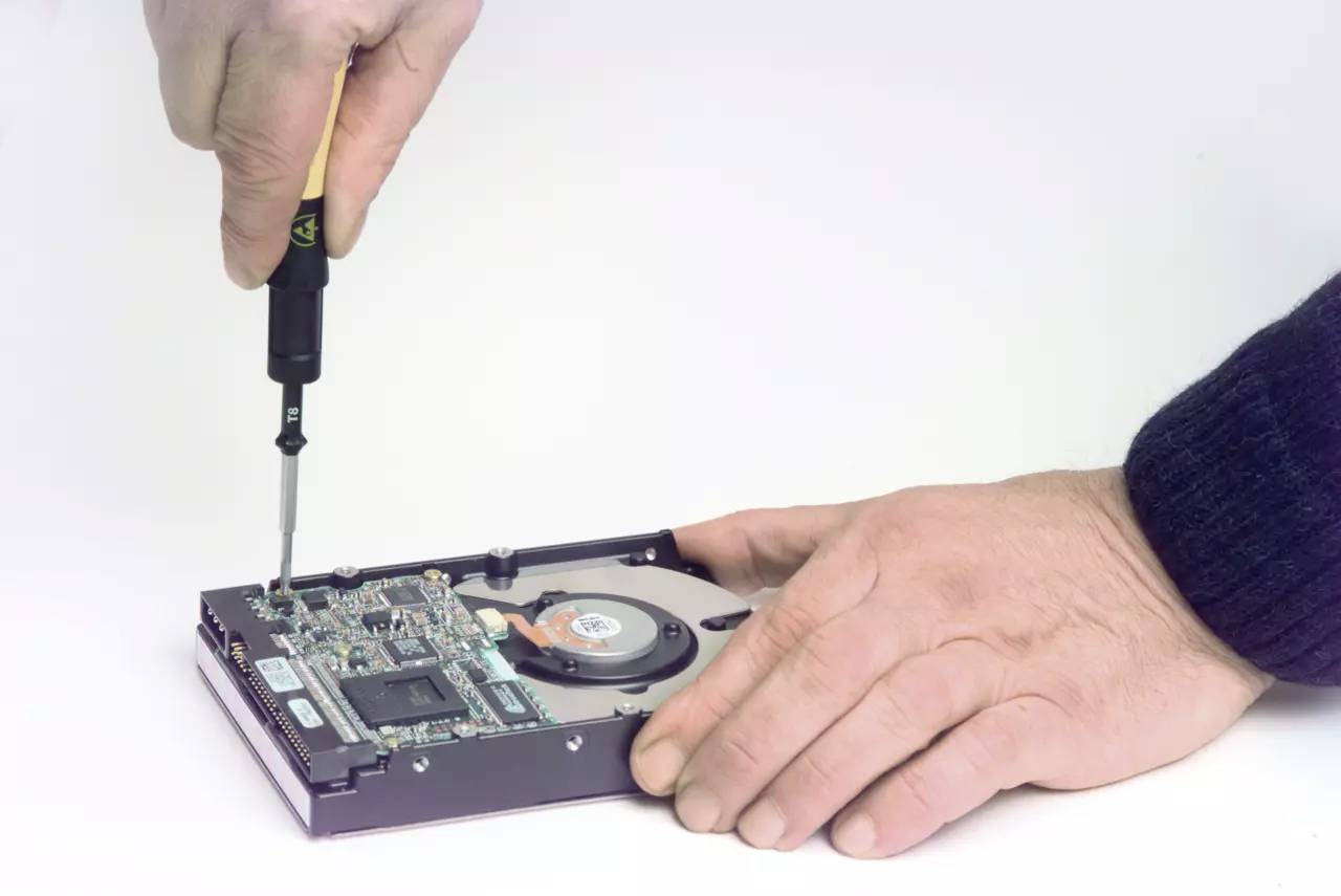 Två händer arbetar med en öppnad hårddisk och en skruvmejsel för att reparera eller underhålla elektroniska komponenter.