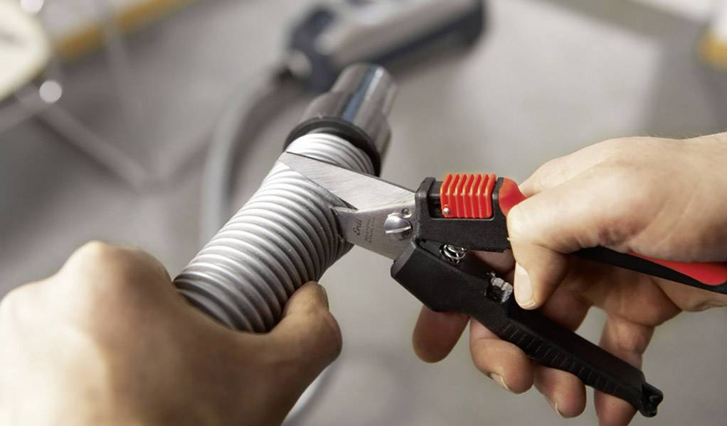 En person klipper ett grått, flexibelt rör med en tång. I bakgrunden syns en oskarp dammsugare.