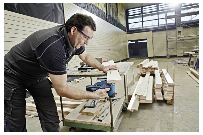 Man använder en elborr för att montera träbräder i en verkstad, med timmer staplade på ett arbetsbord och golv i närheten.