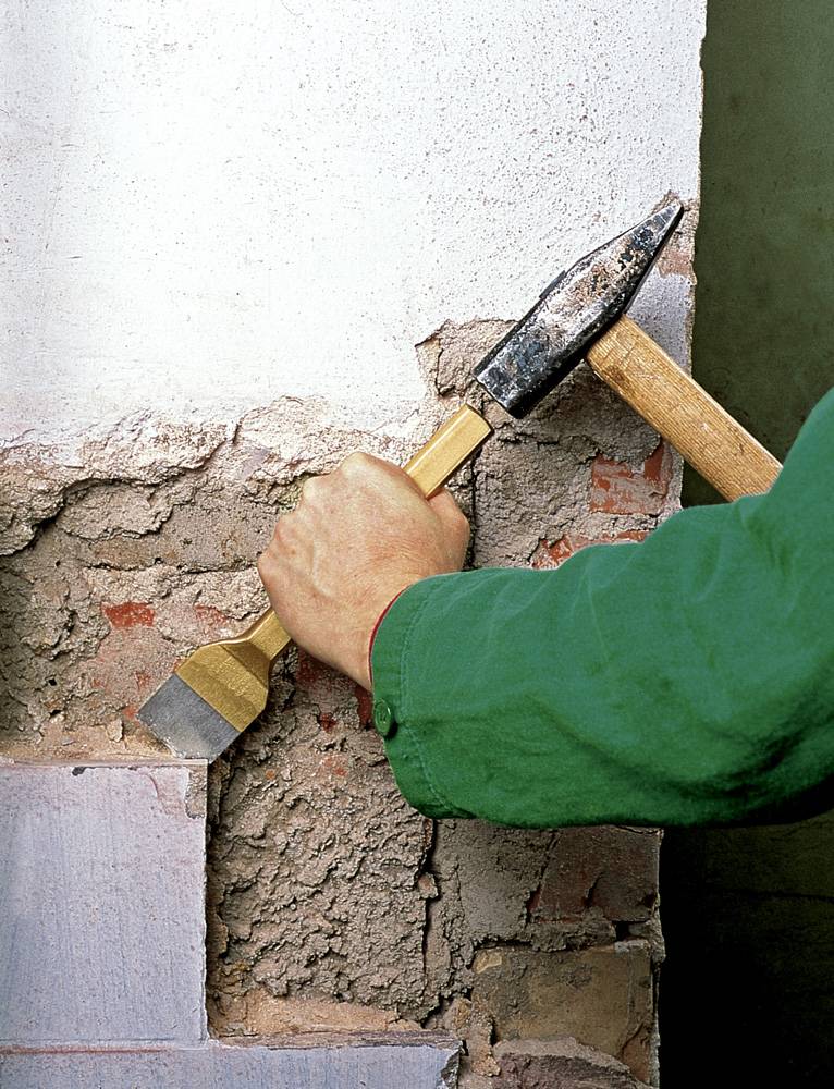 En hantverkare bär en grön overall. Han använder hammare och mejsel för att lossa puts från en mur.