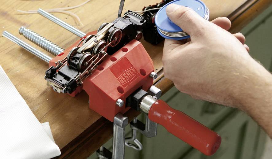 En hand smörjer en roterande metallmekanism som är fastklämd i ett rött skruvstäd med hjälp av en liten blå behållare.