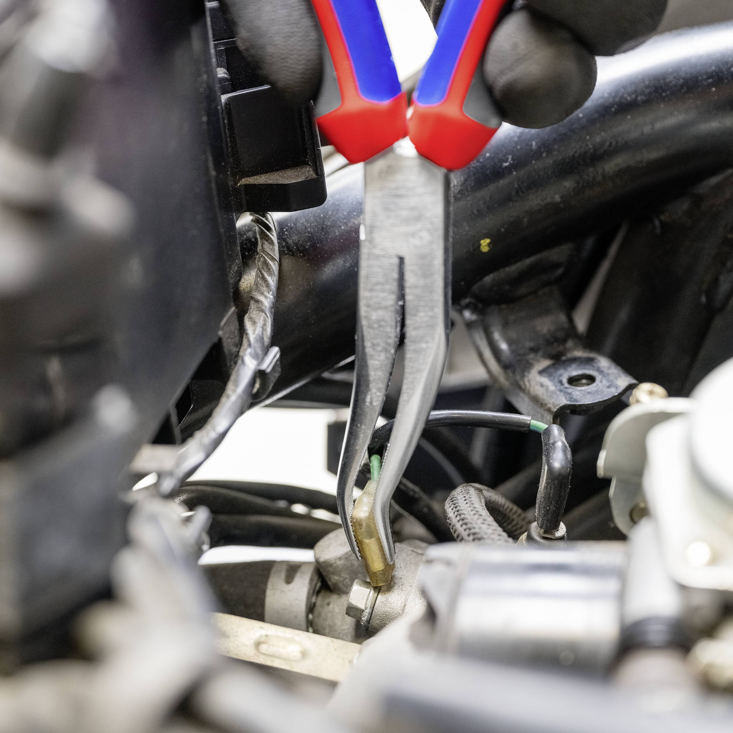 En mekaniker använder en tång för att arbeta med en elektrisk kabel i en motorrum. Kablar och komponenter är synliga.