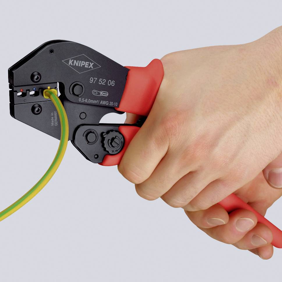 En hand håller fast en krympningstång med röd handtag som pressar en gul-grön kabel.