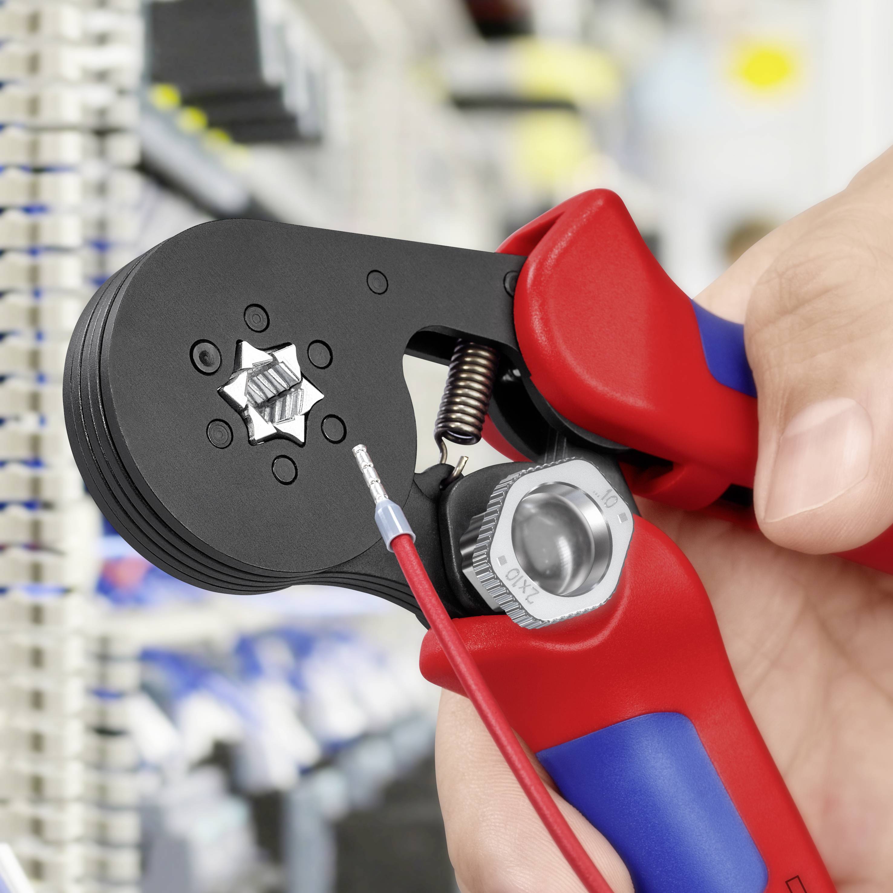 En hand håller en röd crimptång och klämer fast en metallkontakt med en kabel i en teknisk laboratorie- eller verkstadsmiljö.