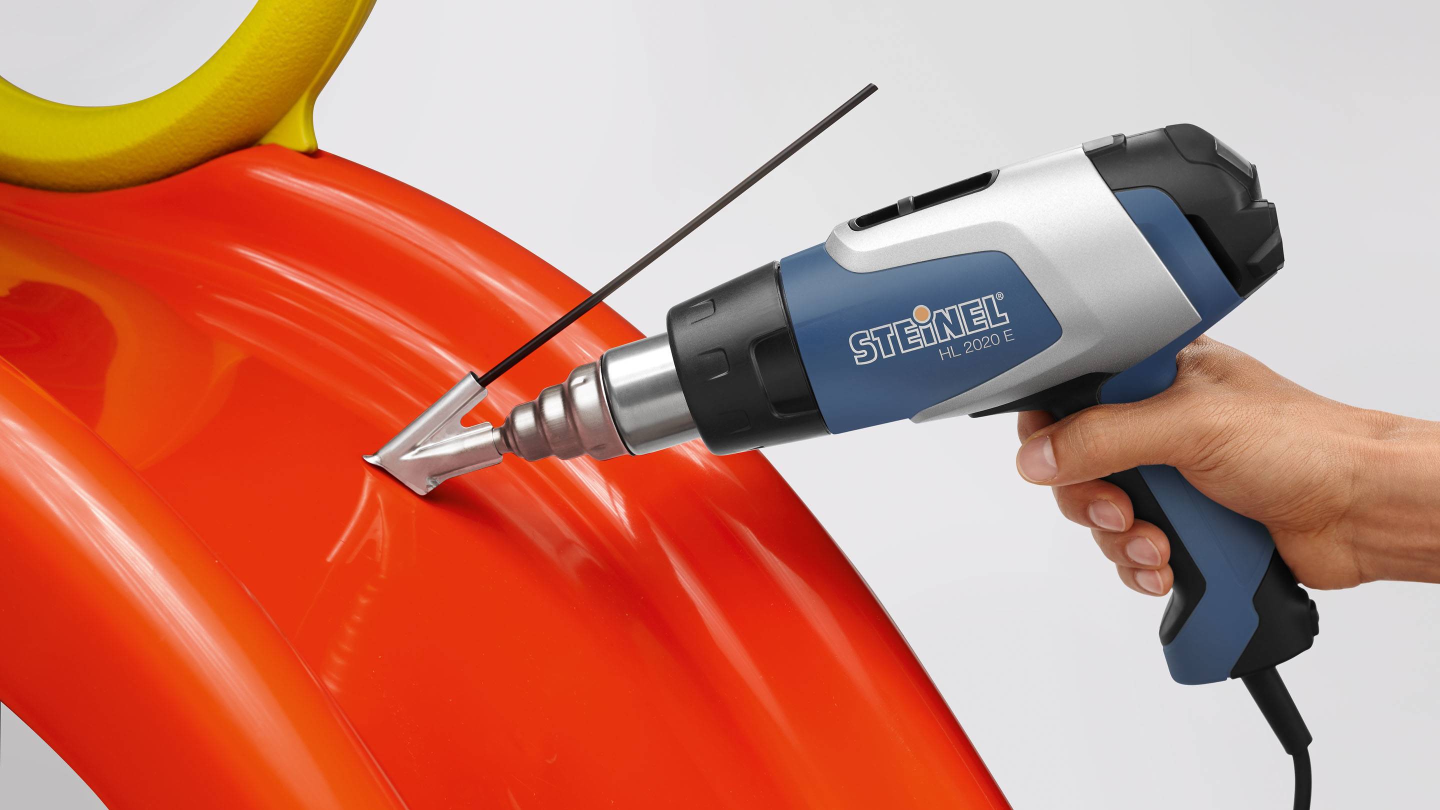 En hand håller ett blå-svart värmepistol som är riktad mot ett stort orangefärgat plastföremål.