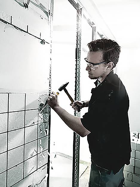 En person som bär skyddsglasögon lägger kakel på en vägg med hjälp av en hammare, vilket betonar precision och byggnadskunskaper.