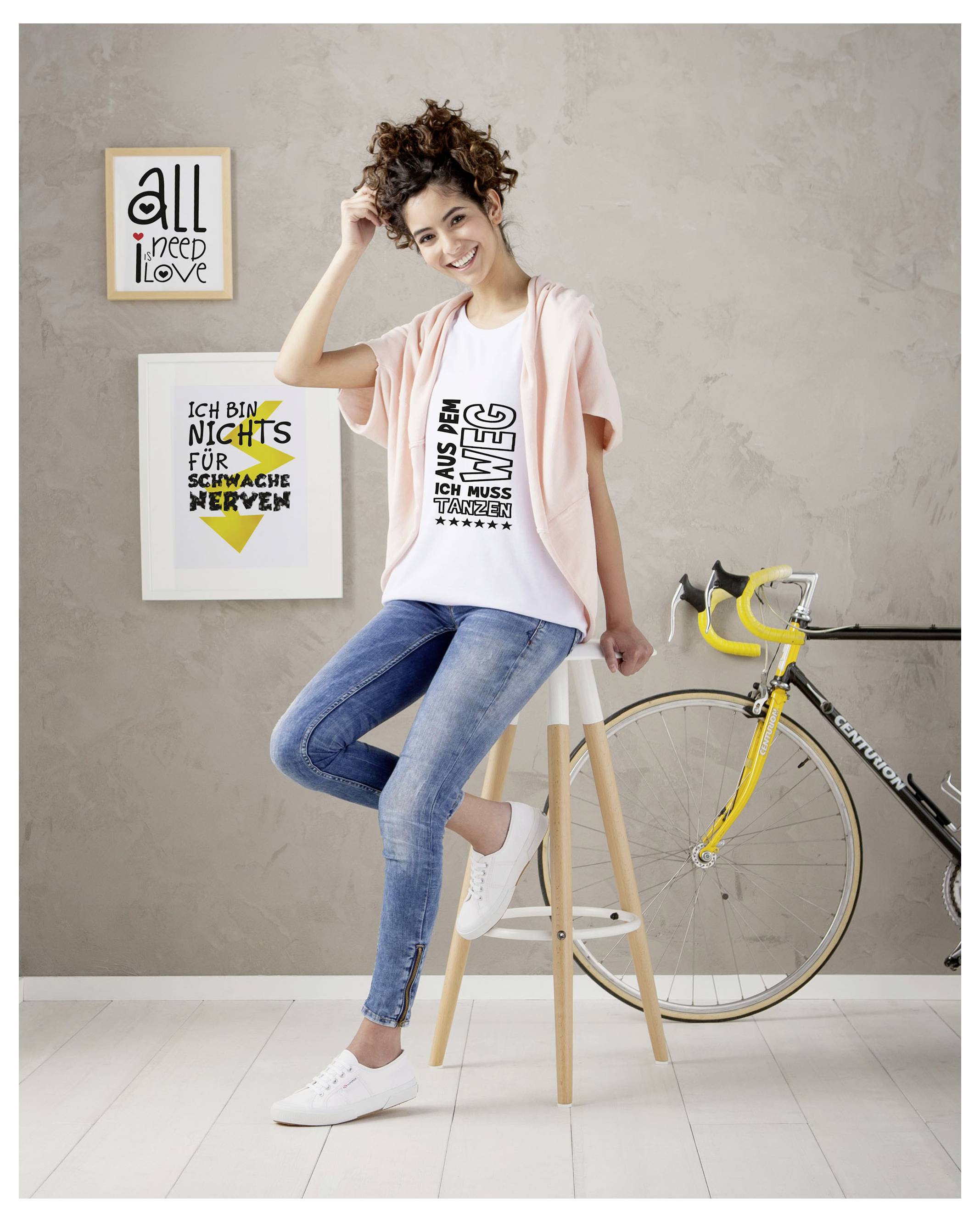 En ung kvinna sitter leende på en pall bredvid en gul tävlingscykel; i bakgrunden hänger två inramade affischer med engelsk och tysk typografi.