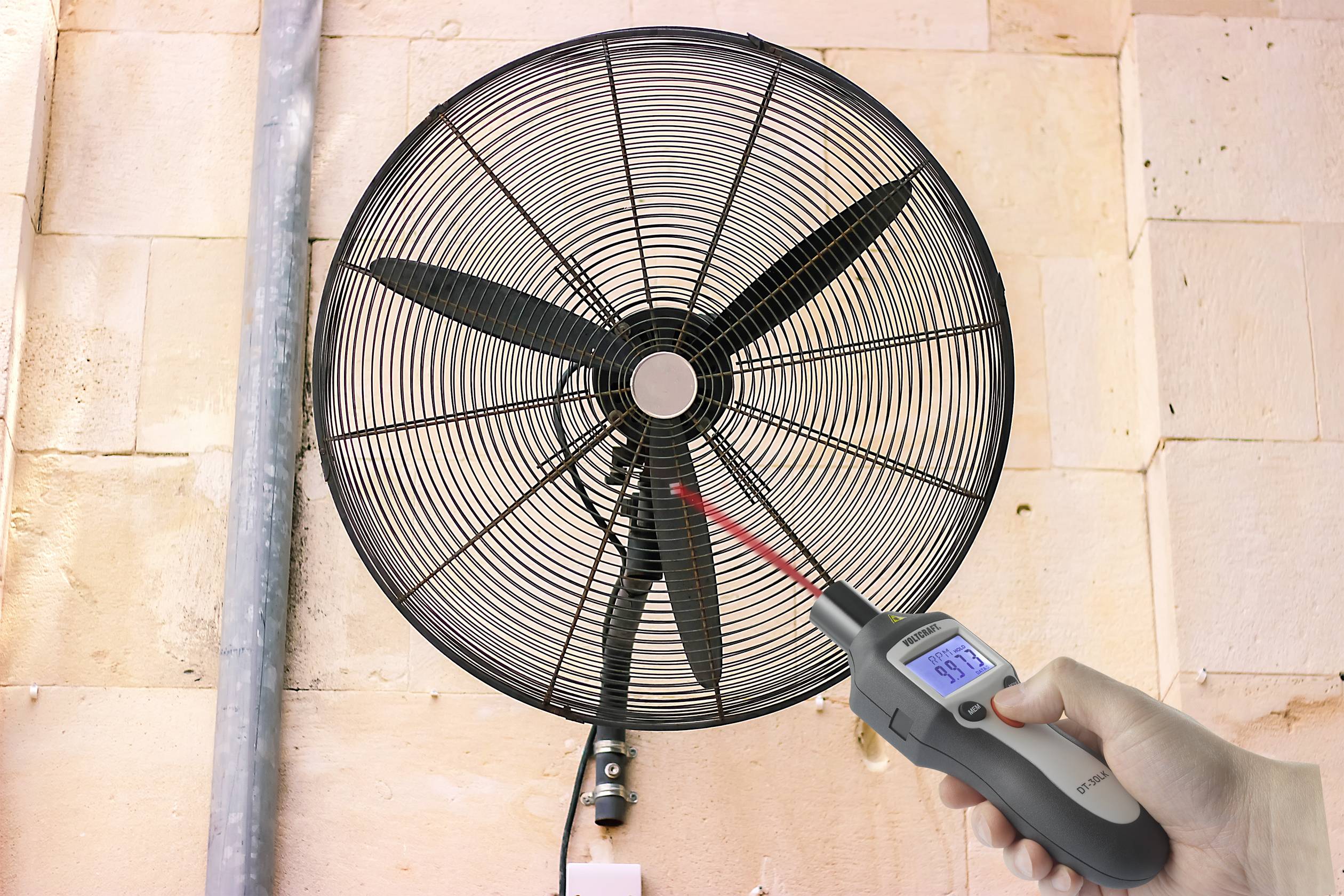 En person mäter vindhastigheten på en stor fläkt med ett digitalt anemometer vid en vägg.