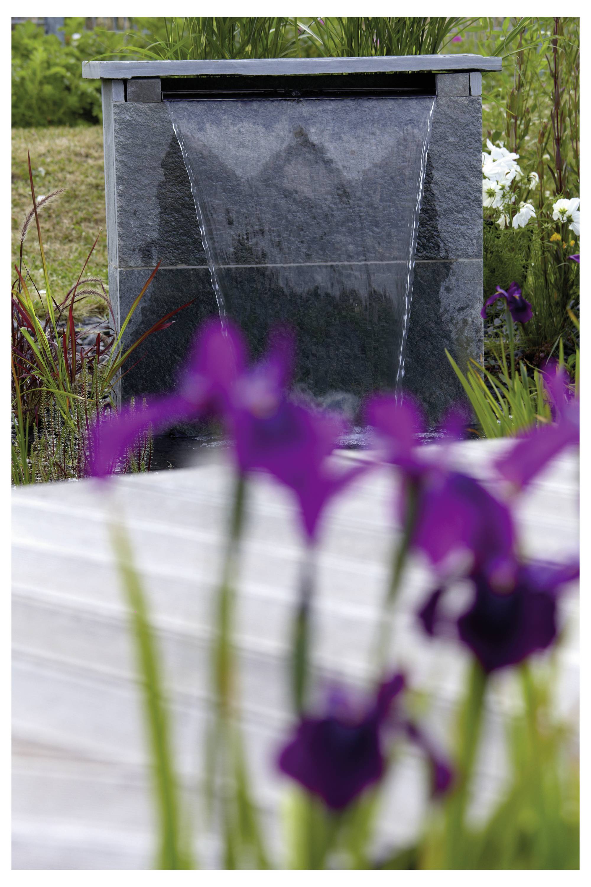 En trädgårdsscen med en stenvatten-installation som låter vatten forsa ner, omgiven av grönska och lila blommor i förgrunden.
