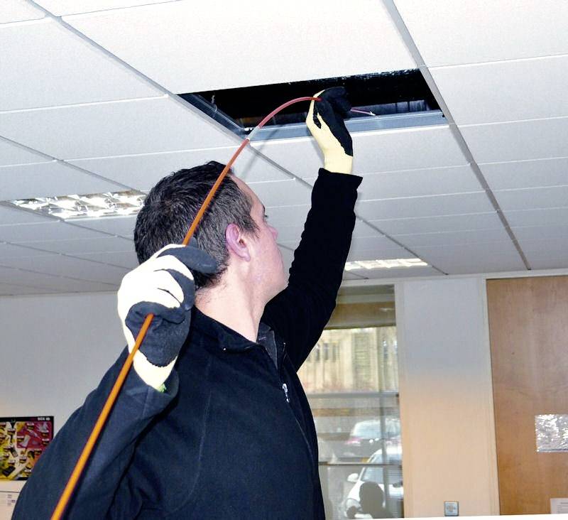 En person i svart klädsel och handskar drar en röd kabel genom en takplatta i en kontorsbyggnad.