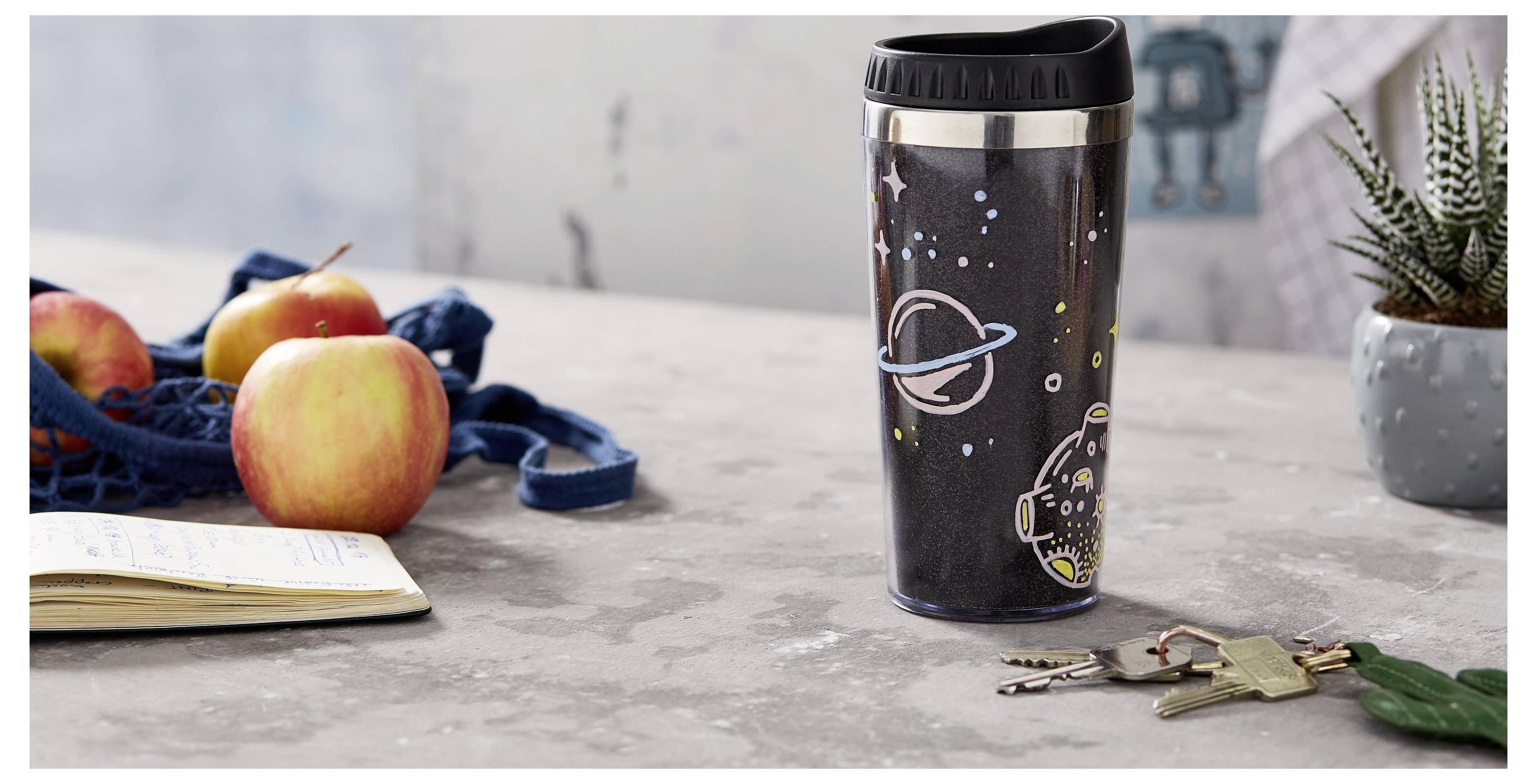 Resemugg med rymdtema på ett bord bredvid äpplen, en anteckningsbok och nycklar, vilket antyder en morgonklar avresa eller en packat lunch-scen.