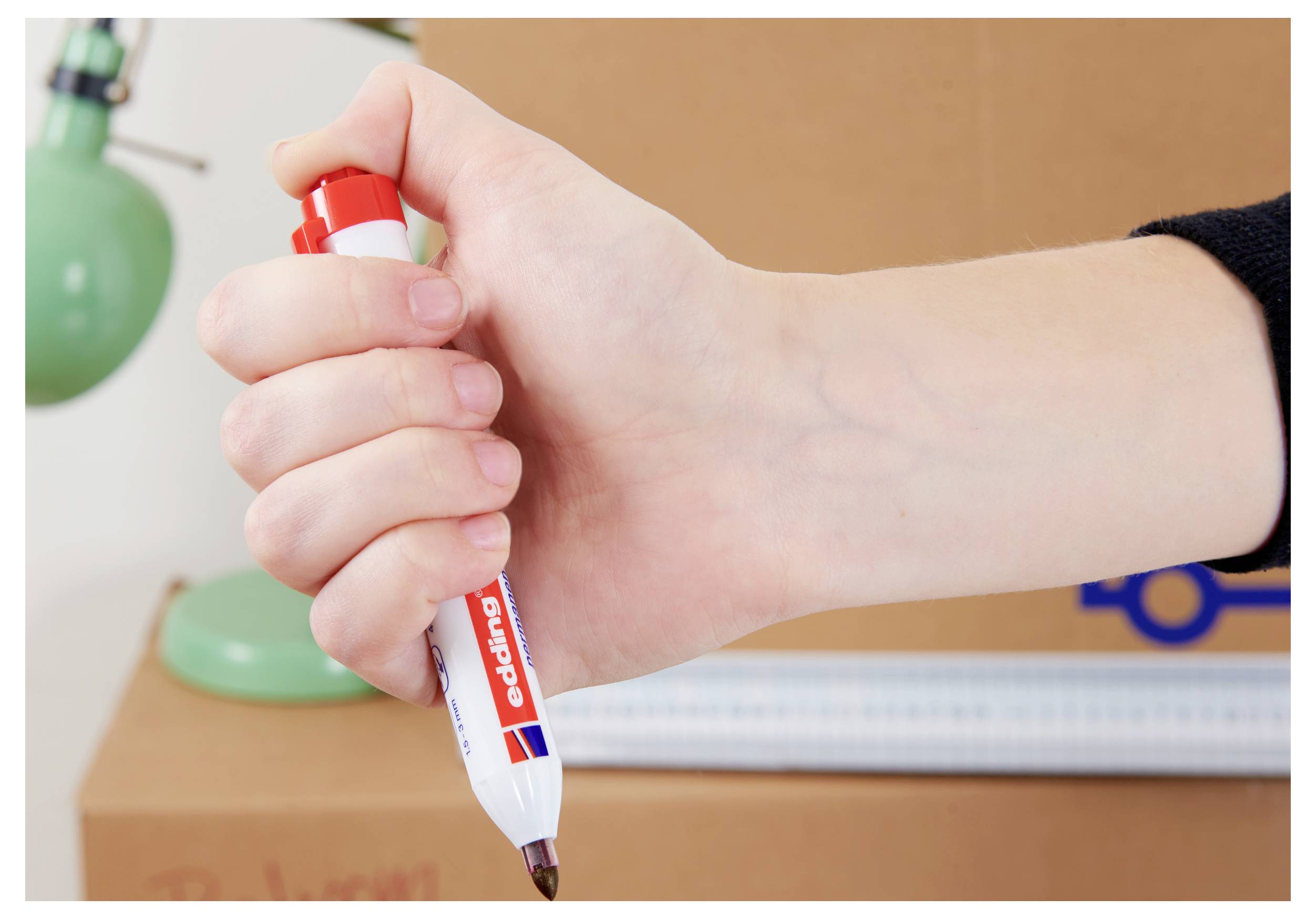 Hand holding a red dry-erase marker poised to write on a whiteboard, with classroom materials blurred in the background.<br><br>Håll med en röd torrbortningstejp markör redo att skriva på en whiteboard, med klassrumsmaterial suddiga i bakgrunden.