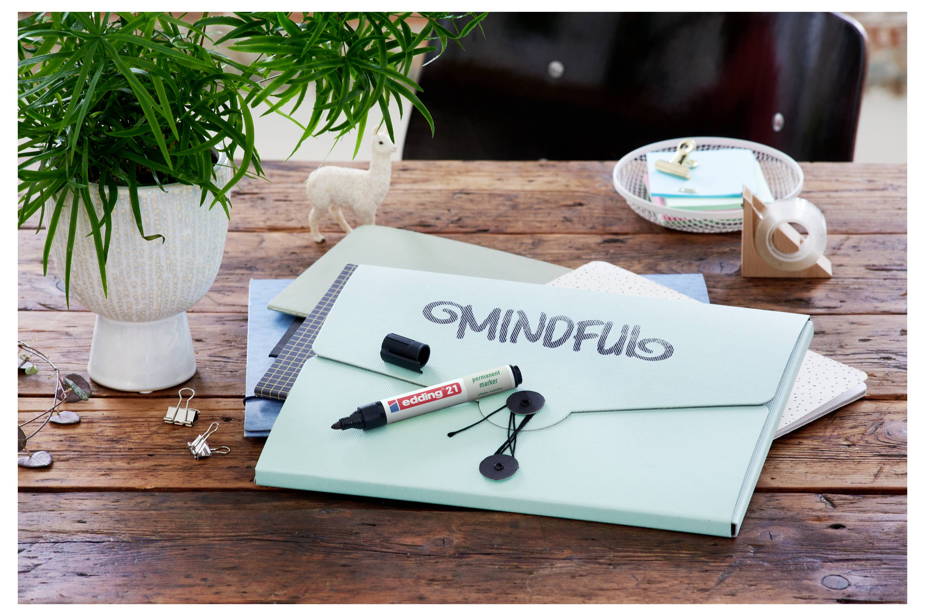 En arbetsutrymmesbild med en mintgrön mapp märkt "Mindful", en markör, öronsnäckor, en anteckningsbok, en växt och skrivbordsföremål på ett trä bord.